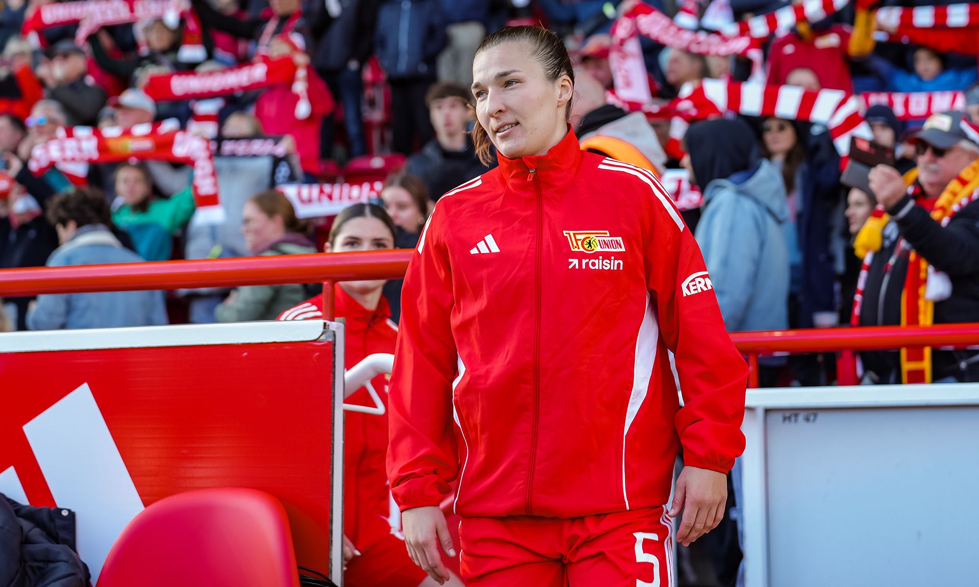 Spielerin in roter Sportjacke betritt das Spielfeld, im Hintergrund Fans mit Schals.