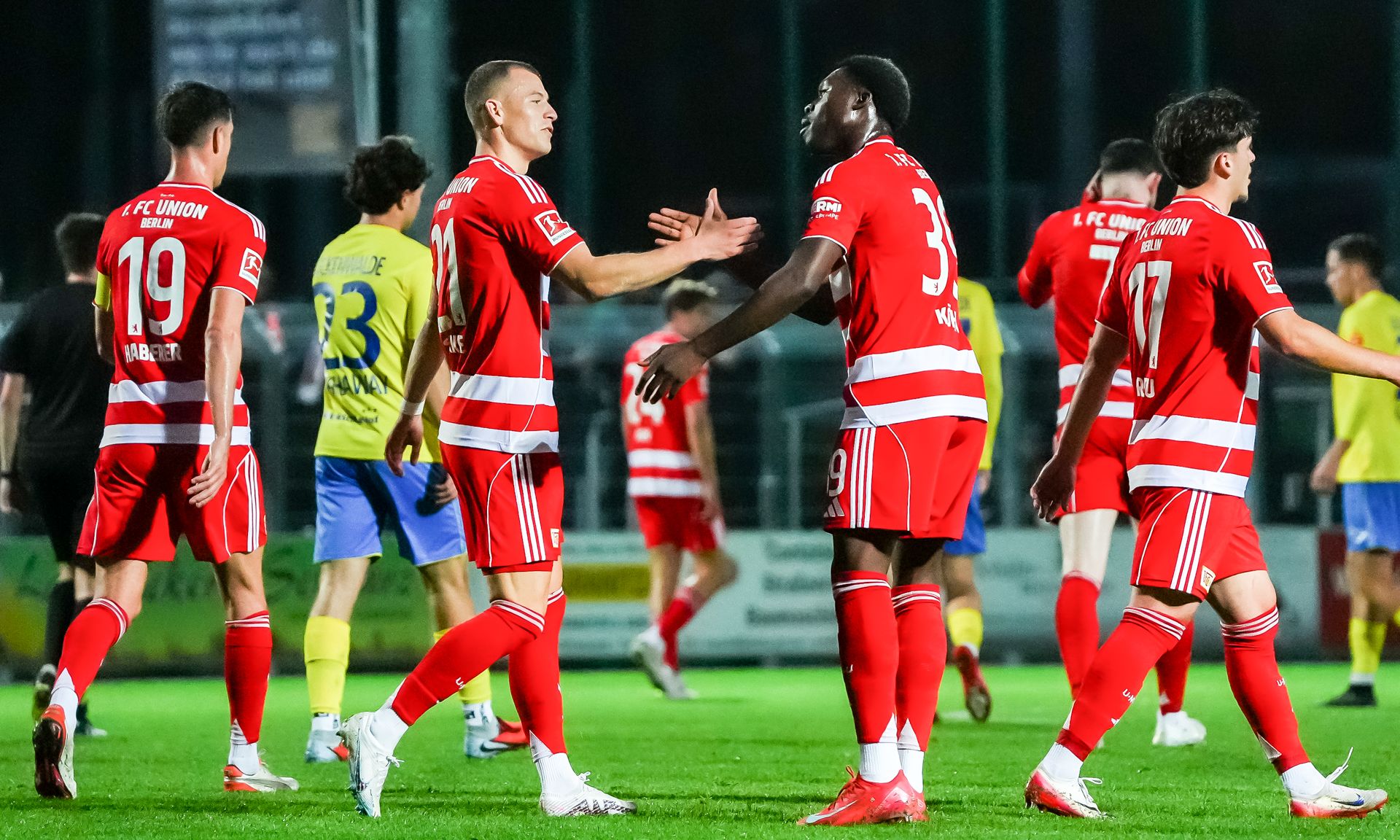 Spieler in roten Trikots feiern nach einem Spiel, während einige Spieler in gelben Trikots im Hintergrund stehen.