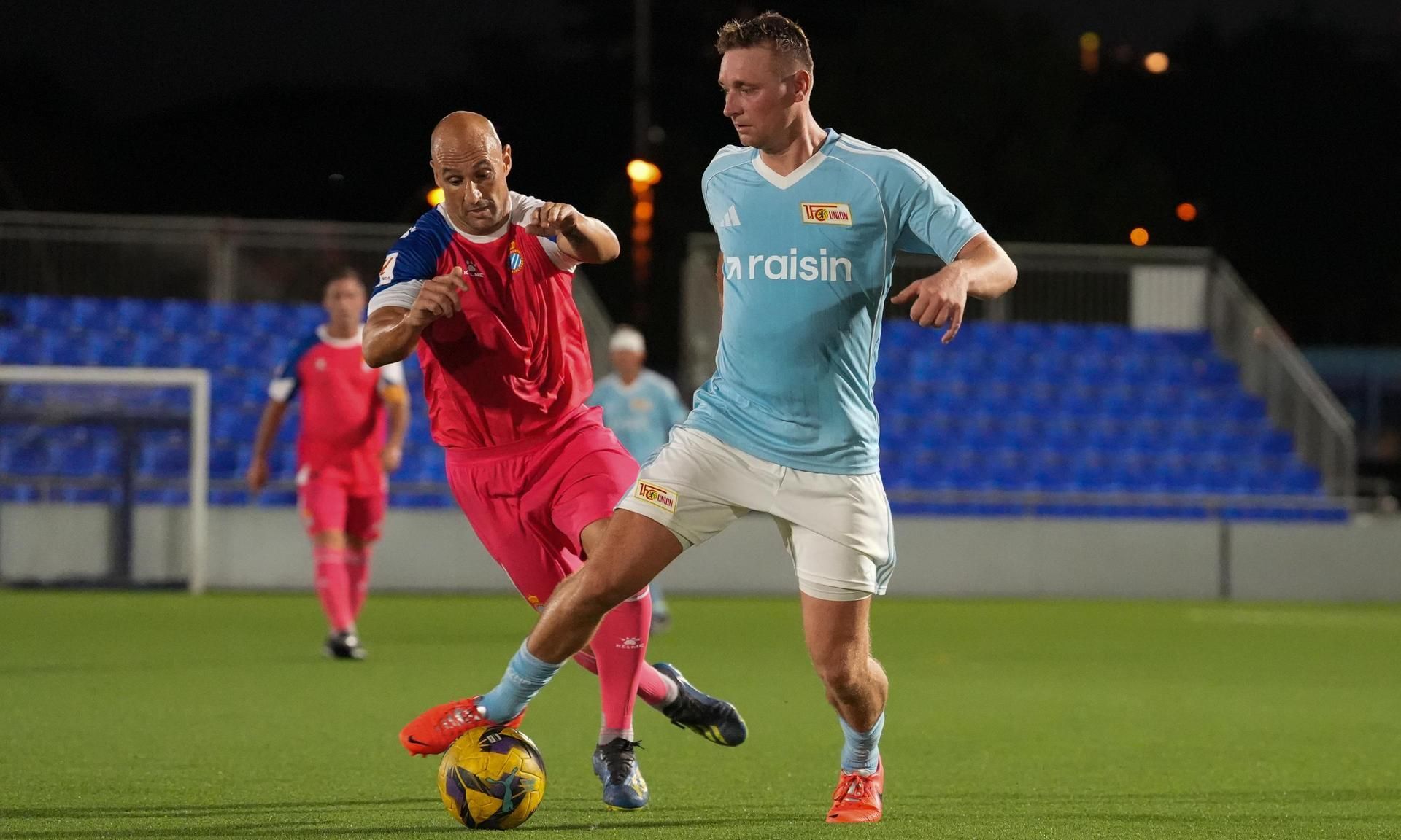 Zwei Fußballspieler kämpfen um den Ball auf einem grünen Platz unter Flutlicht.