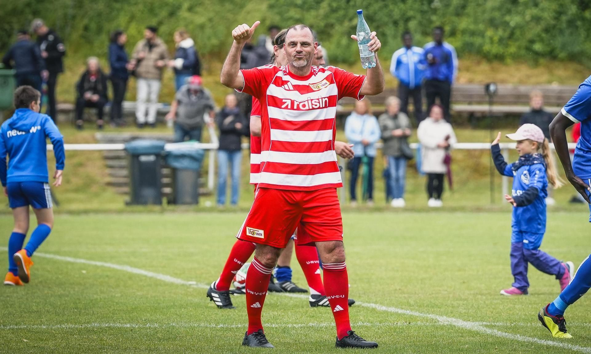 Ein Fußballspieler in einem rot-weißen Trikot hebt die Daumen und hält eine Wasserflasche, während Zuschauer im Hintergrund stehen.