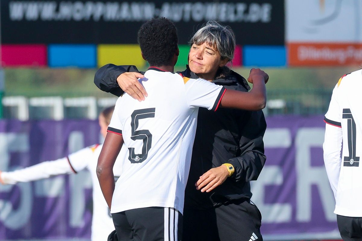Zwei Personen in Sportkleidung umarmen sich auf einem Fußballplatz, eine Trainerin und eine Spielerin mit der Nummer 5.