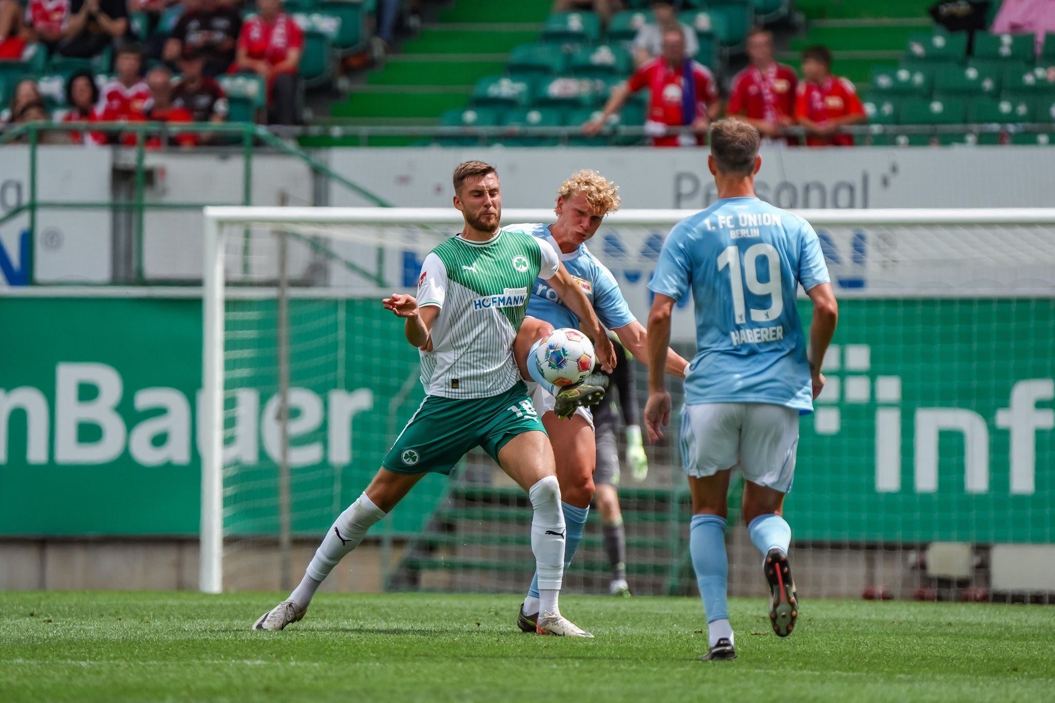 Fußballspieler in grünen und blauen Trikots während eines Spiels, einer passt den Ball, Zuschauer auf der Tribüne im Hintergrund.
