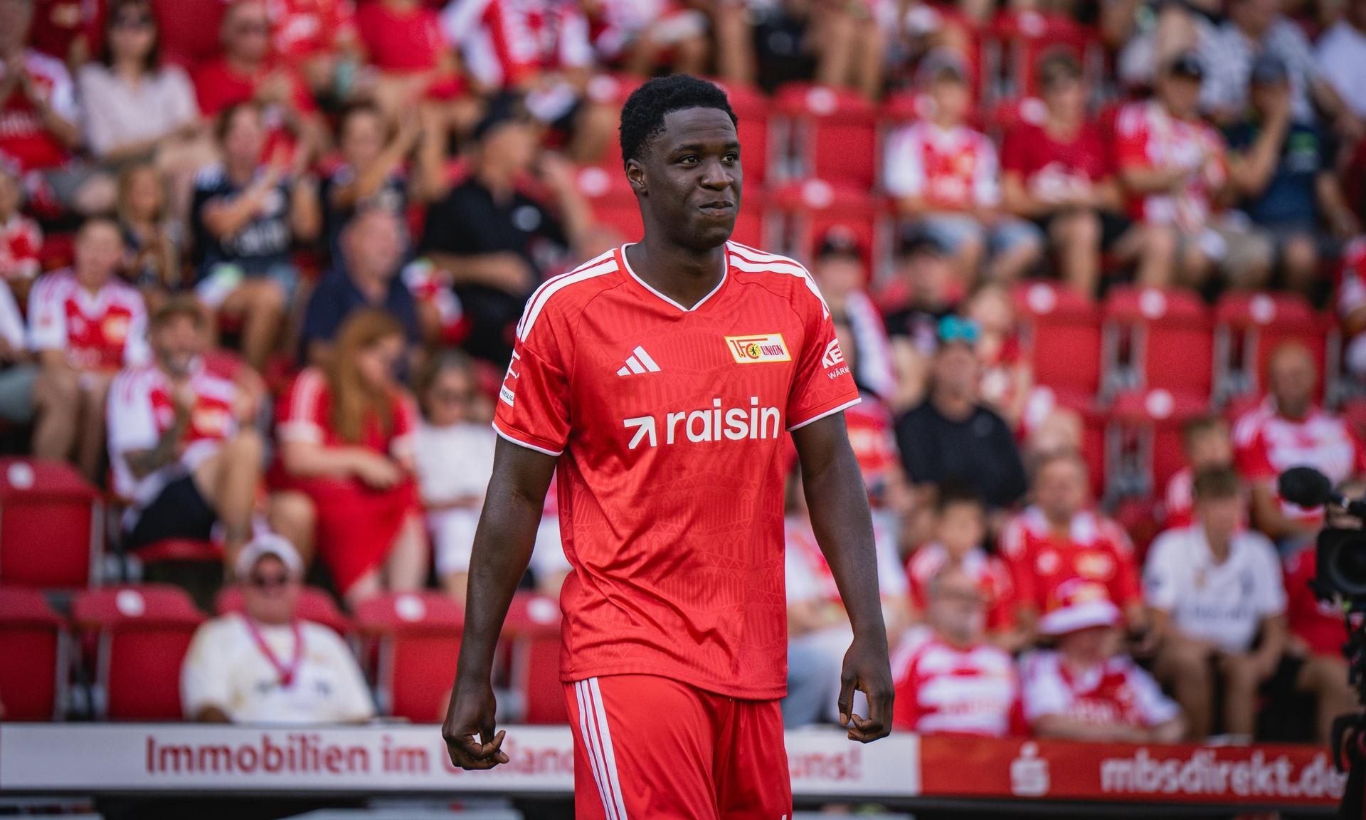 Fußballspieler in rotem Trikot steht auf dem Feld, von Zuschauern in rot-weißen Farben umgeben.