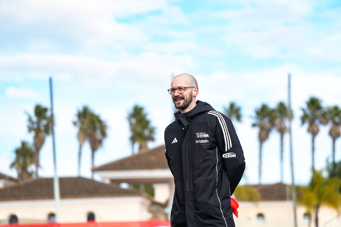Ein lächelnder Trainer in schwarzer Sportkleidung steht auf einem Trainingsfeld, umgeben von Palmen und einem klaren Himmel.