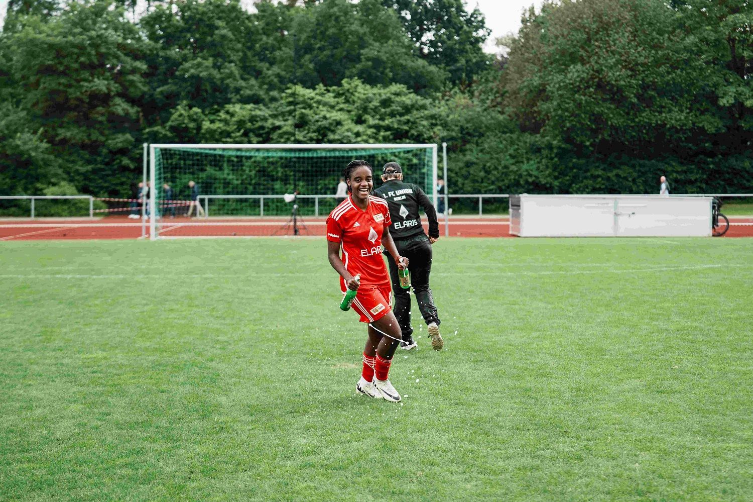 Eine Fußballspielerin in rotem Trikot lächelt und läuft auf einem Rasenplatz, während ein Kollege im Hintergrund zu sehen ist.