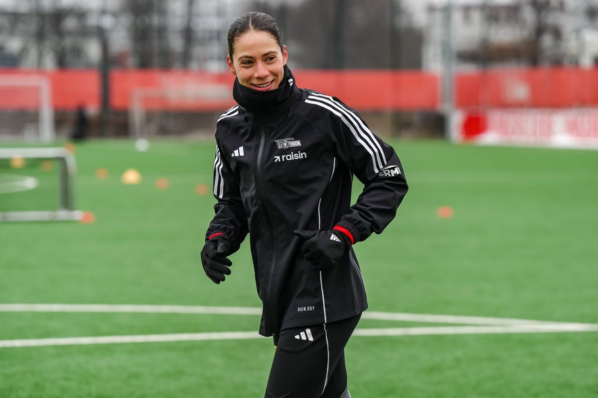 Eine Fußballspielerin in Trainingskleidung lächelt auf dem Platz, umgeben von Trainingsmaterialien.