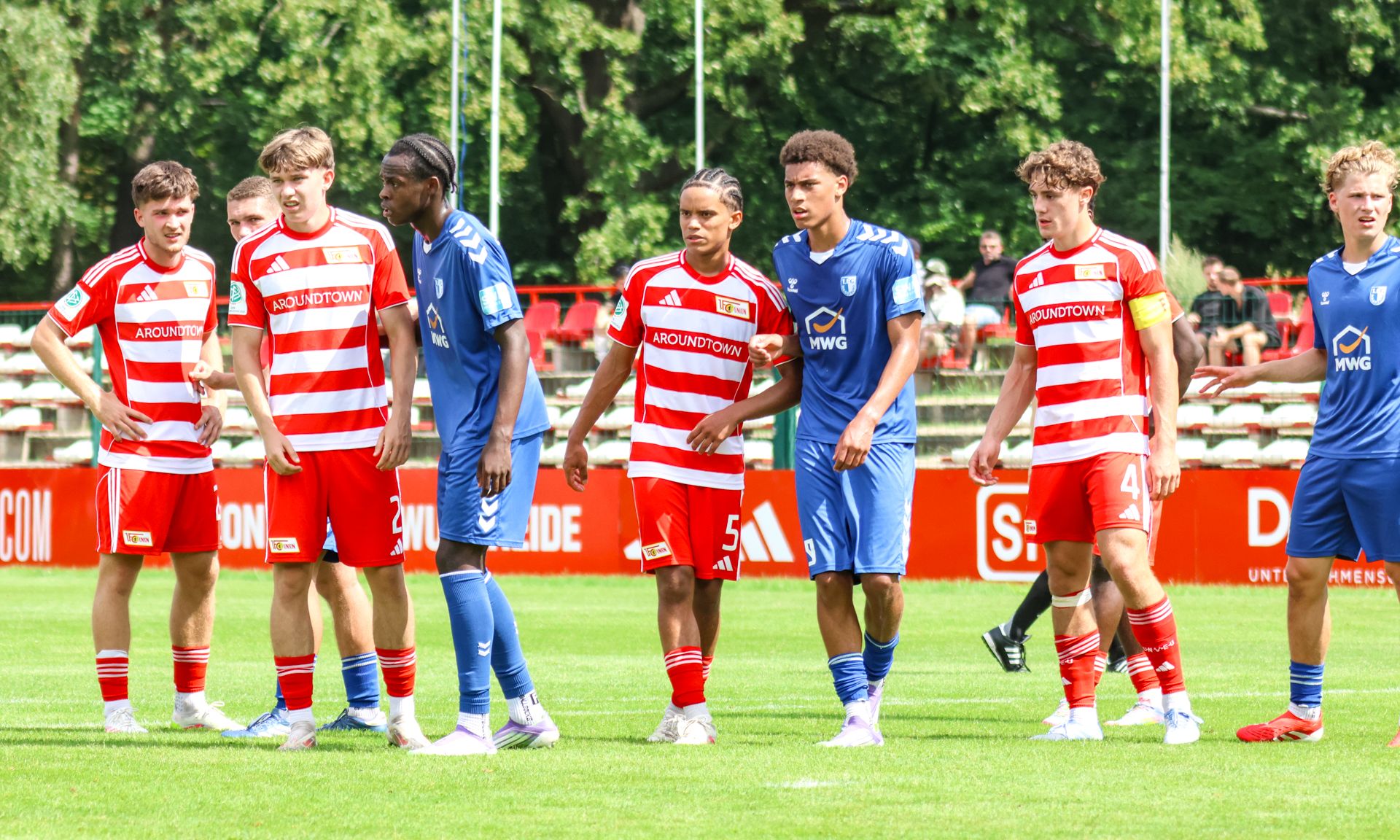 Spielszene mit Fußballspielern in roten und blauen Trikots auf einem Rasenfeld, umgeben von Bäumen und Zuschauern.