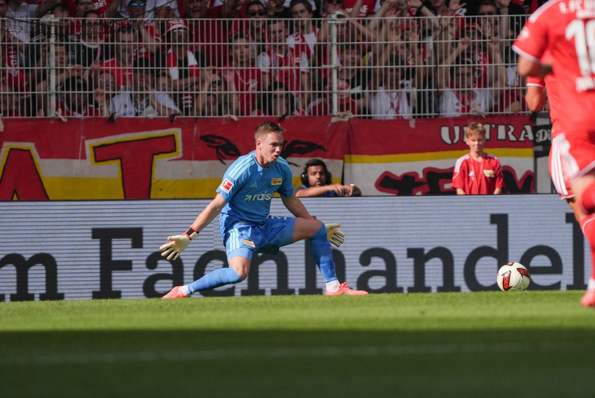 Torwart in blauer Ausrüstung während eines Fußballspiels, der sich nach einem Schuss streckt. Im Hintergrund jubelnde Fans.