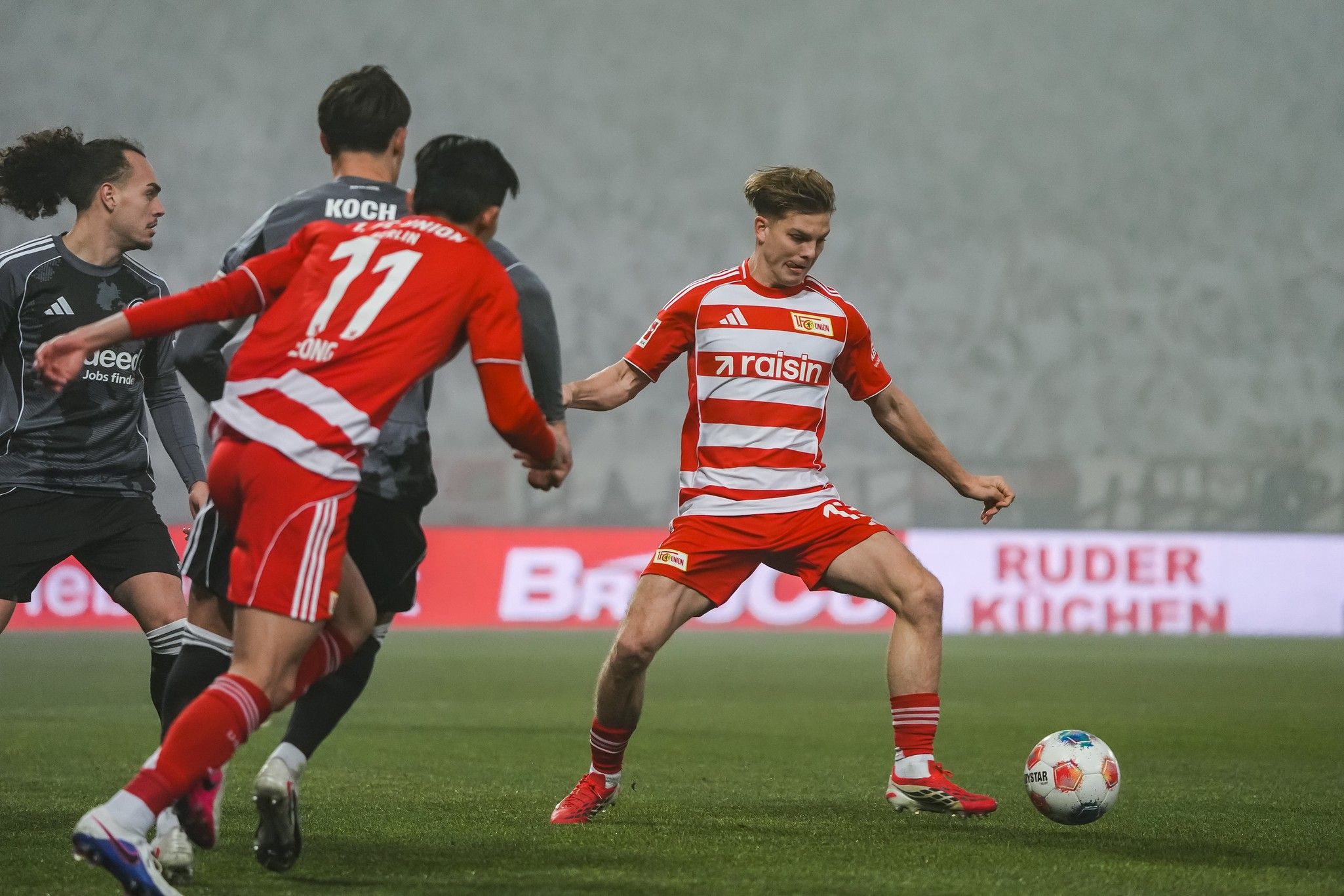 Ein Fußballspieler in einem roten und weißen Trikot tritt den Ball, während zwei Mitspieler in dunklen Trikots sich bewegen.