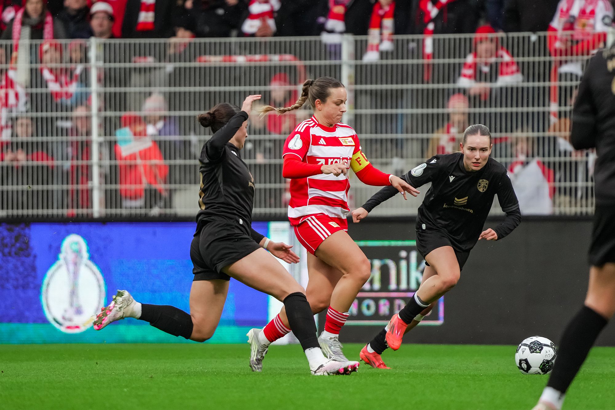 Eine Fußballspielerin in rotem Trikot dribbelt den Ball, während zwei gegnerische Spielerinnen versuchen, sie zu stoppen.