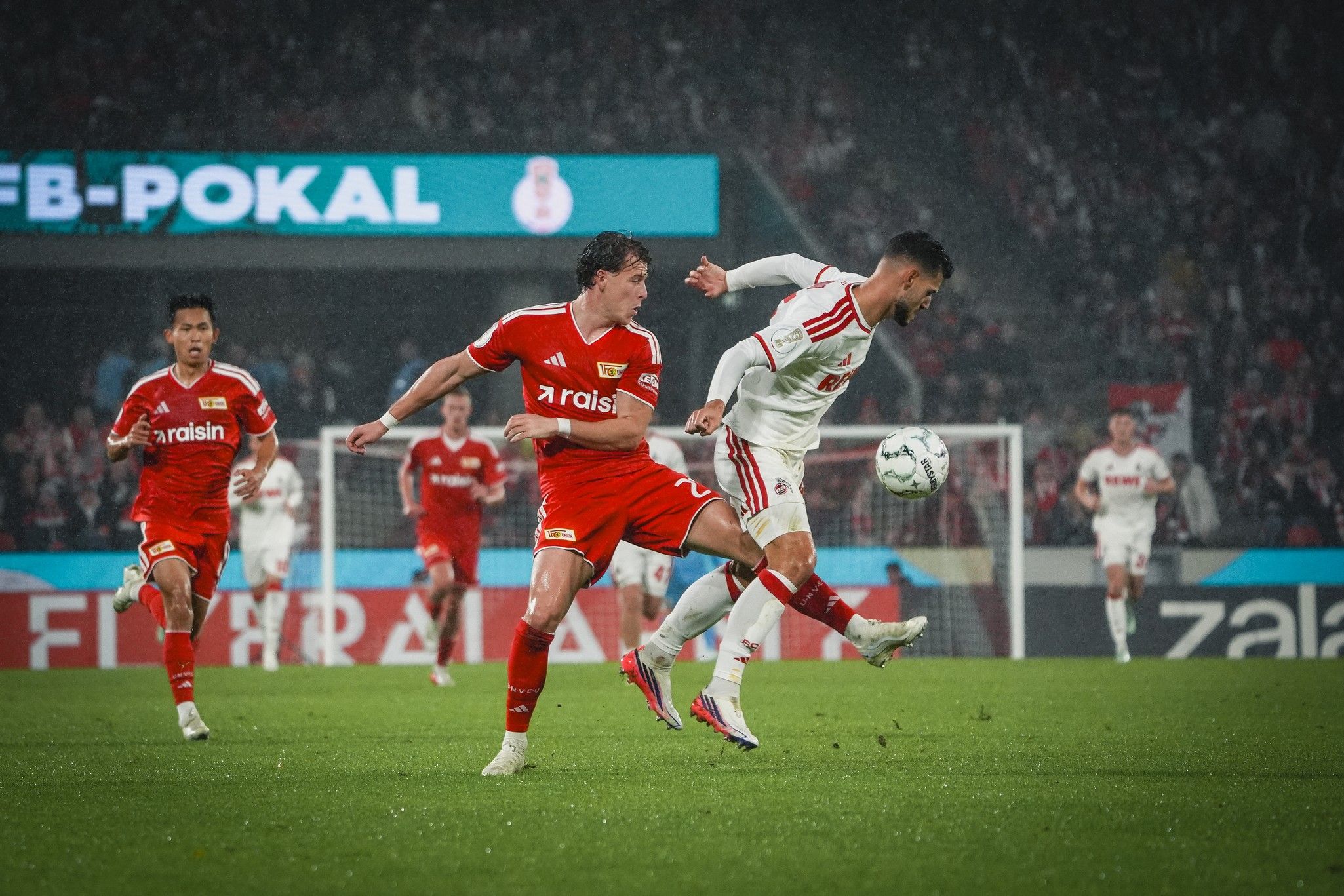 Spielszene im Fußball: Spieler in roten und weißen Trikots kämpfen um den Ball auf einem grünen Spielfeld.