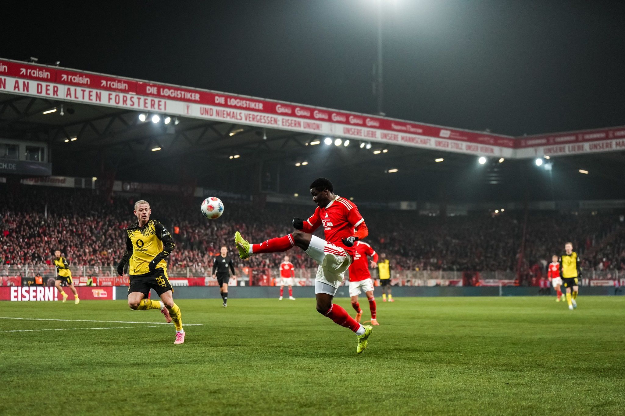 Fußballspiel bei Nacht, Spieler in roter Kleidung steht im Fokus, seine Bewegung zeigt einen Pass, Fans im Hintergrund.
