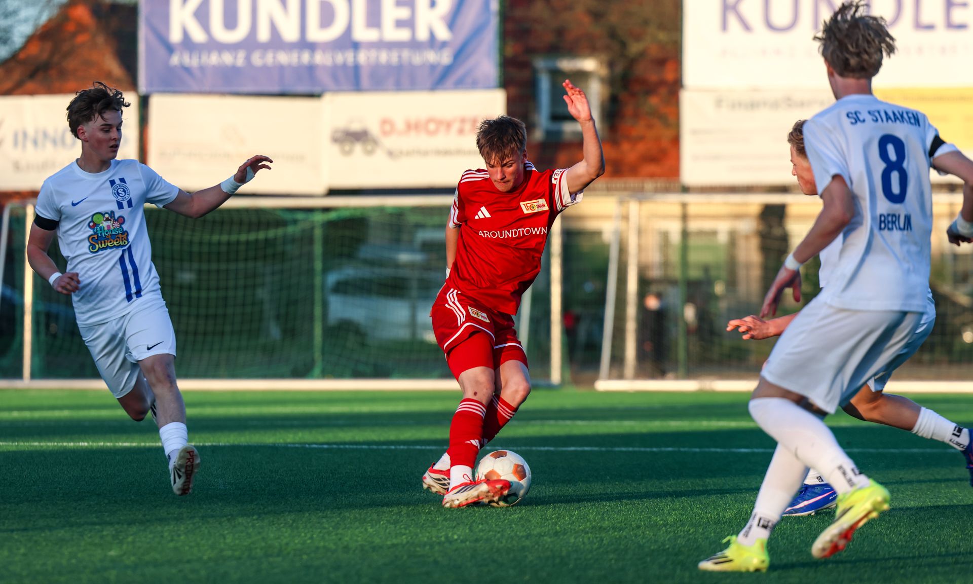 Ein Fußballspieler in roter Trikott färbt während eines Spiels auf einem Kunstrasenplatz. Zwei Spieler in blauen Trikots sind in der Nähe.