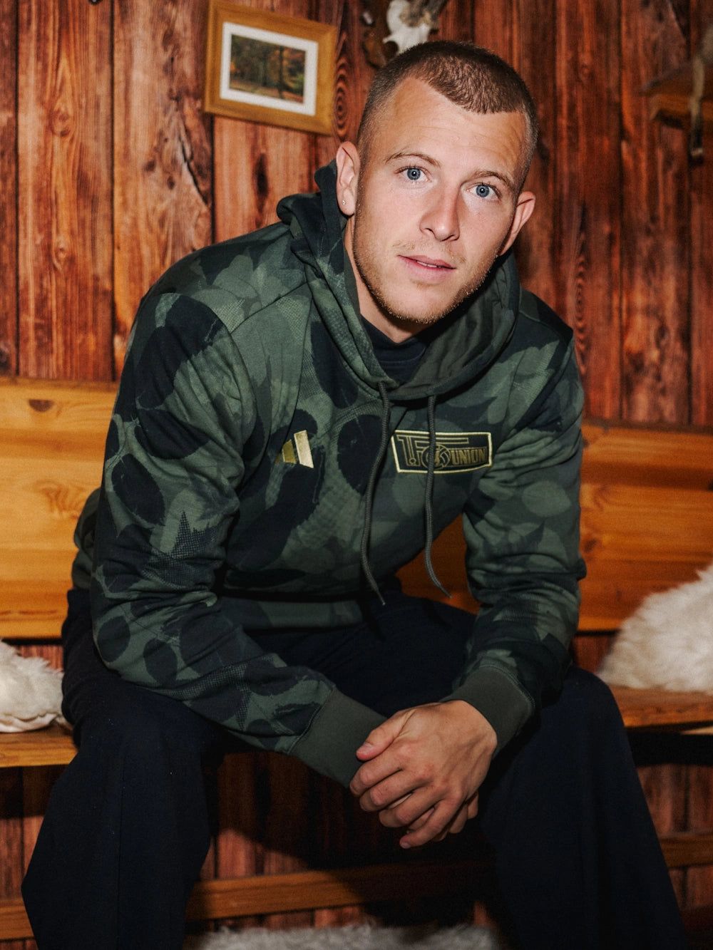 Mann im Camouflage-Hoodie sitzt auf einer Holzbank vor einer rustikalen Holzverkleidung.