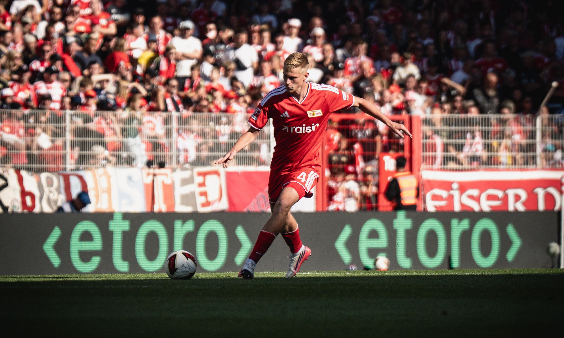 Ein Fußballspieler in rotem Trikot dribbelt den Ball auf dem Platz, umgeben von jubelnden Fans im Hintergrund.