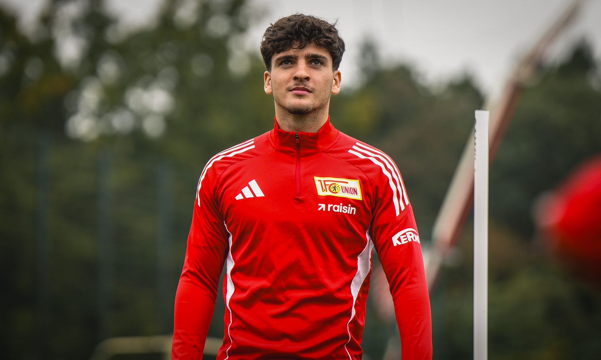 Ein junger Fußballspieler in roter Trainingskleidung steht auf einem Sportplatz, Hintergrund mit Bäumen und verschwommenen Spielern.