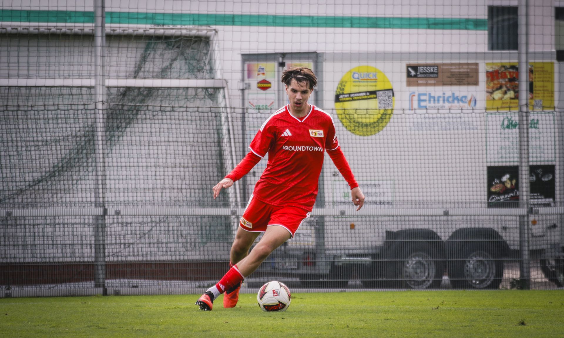 Ein junger Fußballspieler in rotem Trikot dribbelt einen Ball auf einem Sportplatz.