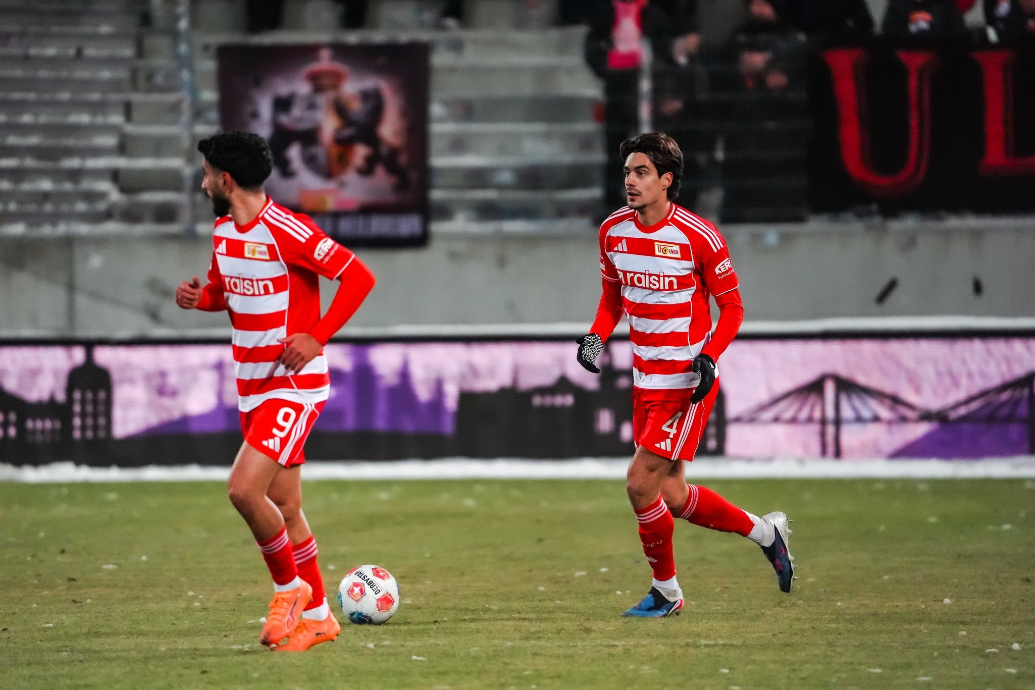 Zwei Fußballspieler in rot-weiß gestreiften Trikots laufen über das Spielfeld. Im Hintergrund sind Zuschauer und Banner zu sehen.