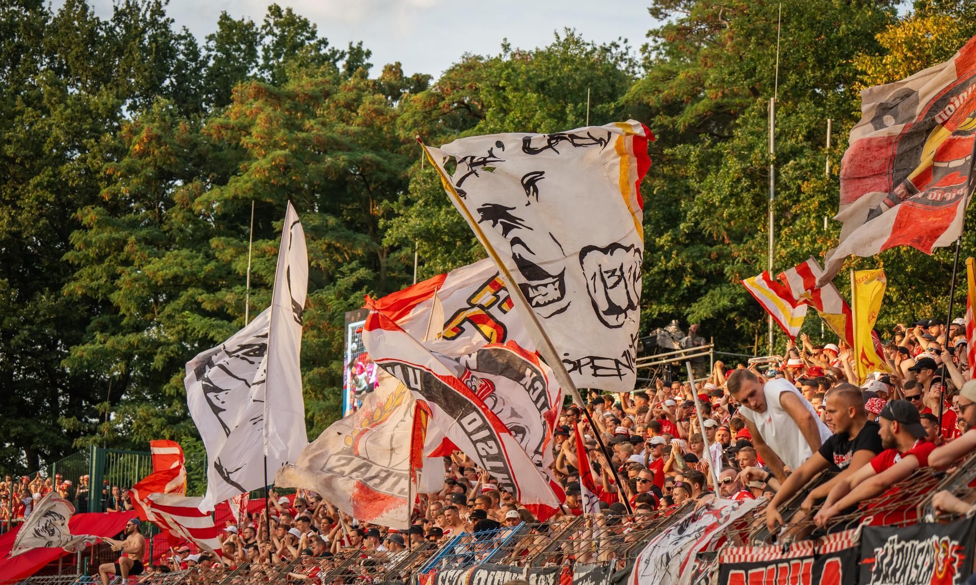 Anfeuernde Fans in einem Stadion, umgeben von bunten Fahnen und Transparenten, mit Bäumen im Hintergrund.