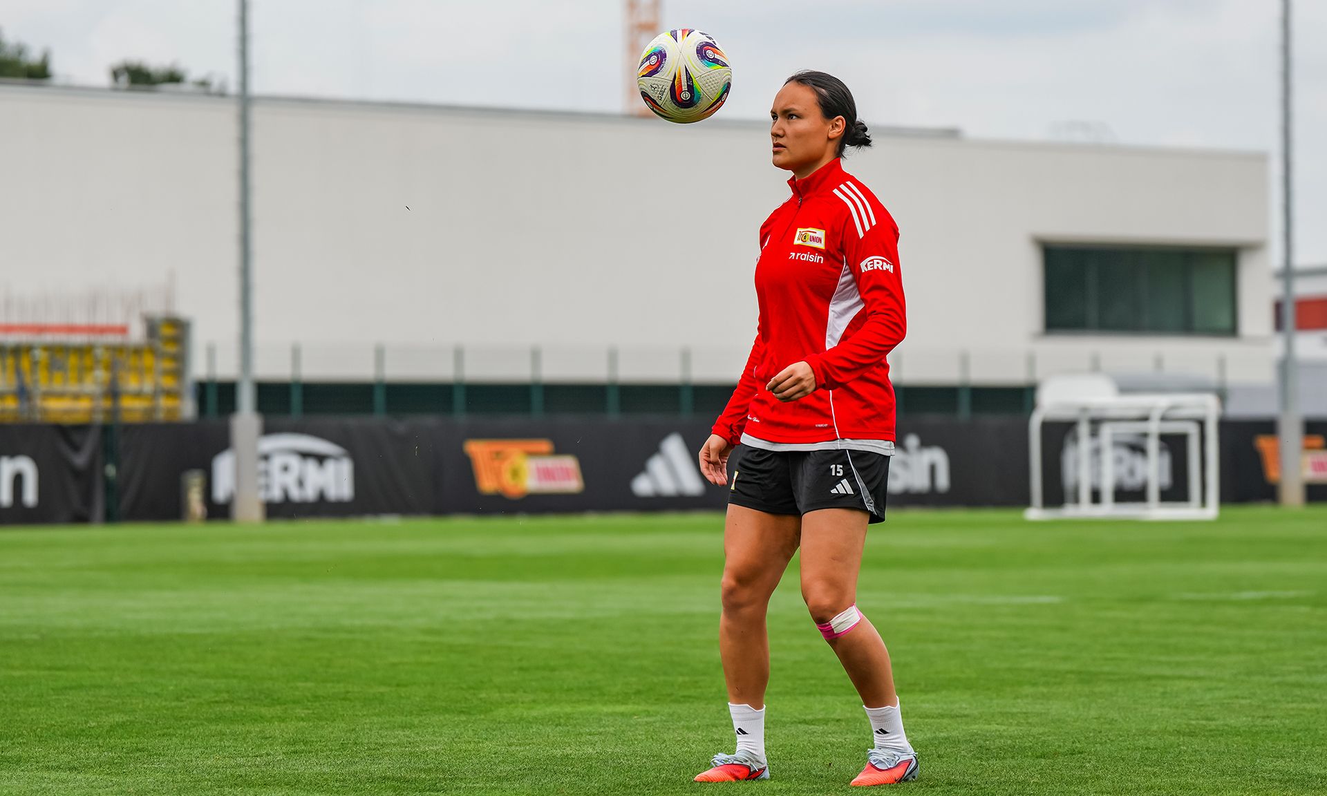 Frau in rotem Sportoutfit jongliert einen Fußball auf einem Trainingsfeld.