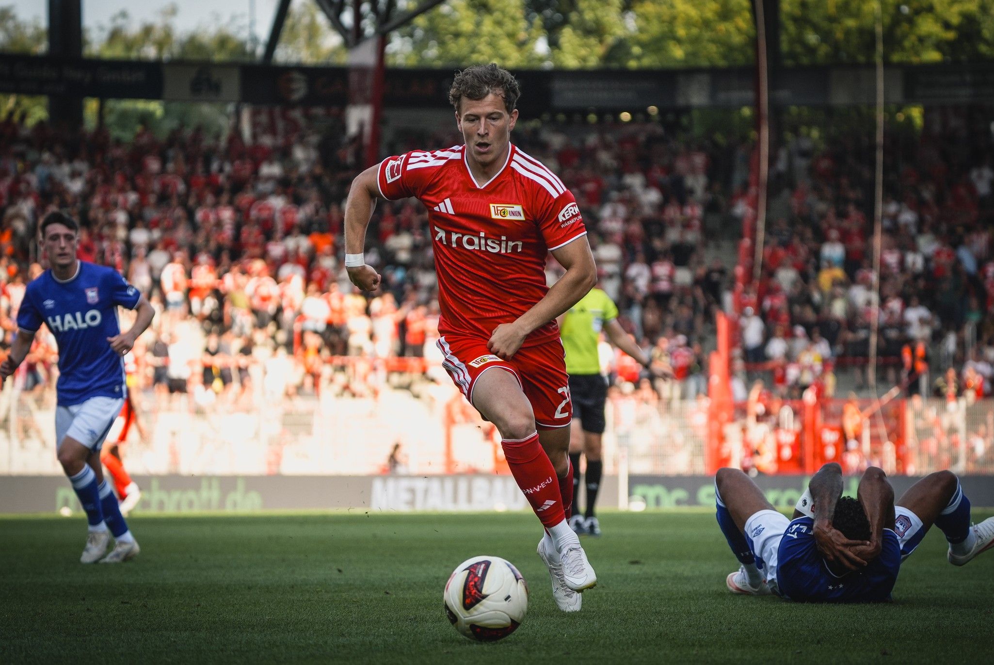 Ein Spieler in rotem Trikot sprintet über das Spielfeld, während ein gegnerischer Spieler am Boden liegt. Zuschauer im Hintergrund.