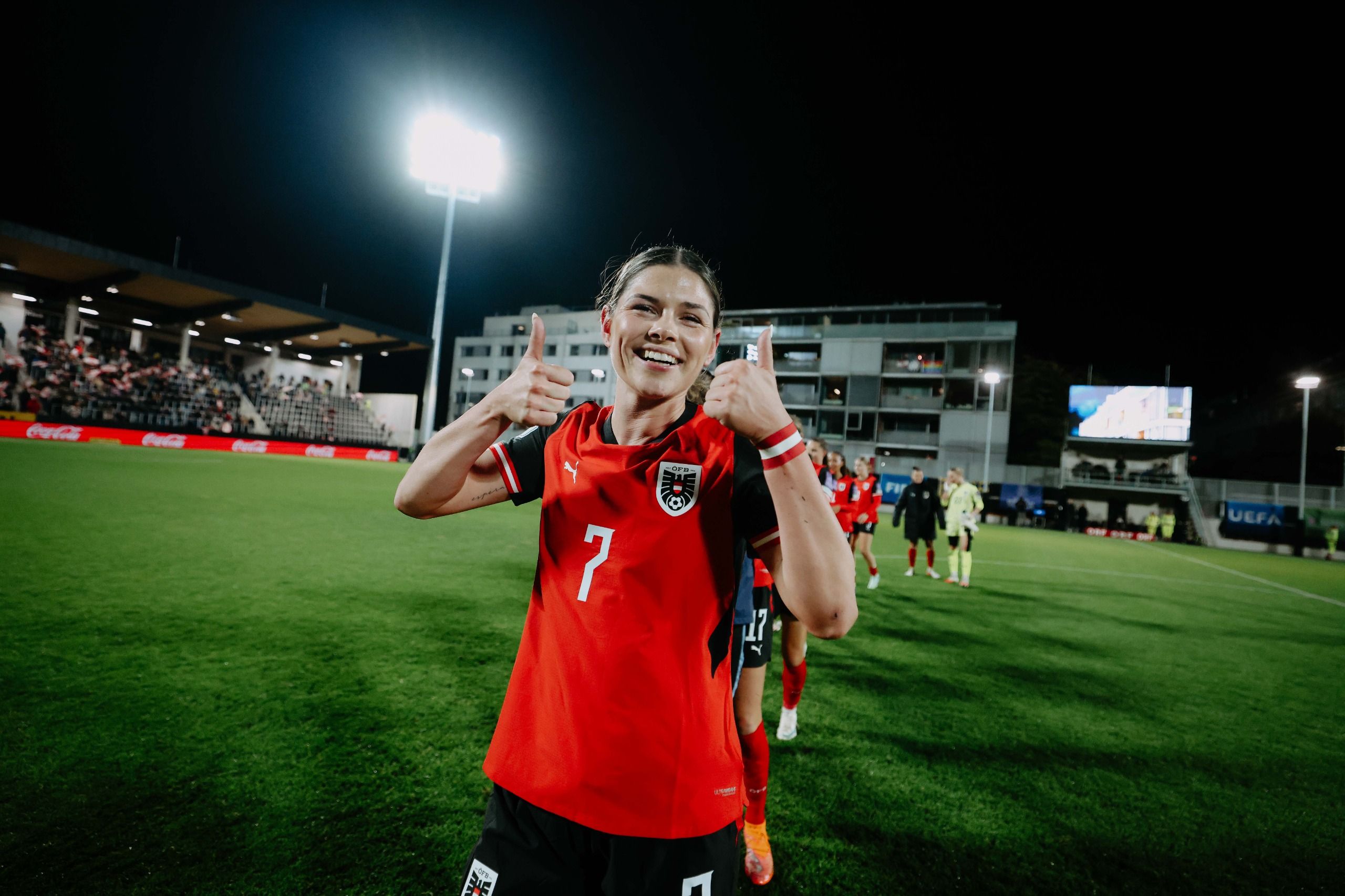 Spielerin in rotem Trikot zeigt Daumen hoch auf dem Fußballfeld bei Nacht, im Hintergrund sind Teamkollegen und Stadioni beleuchtet.