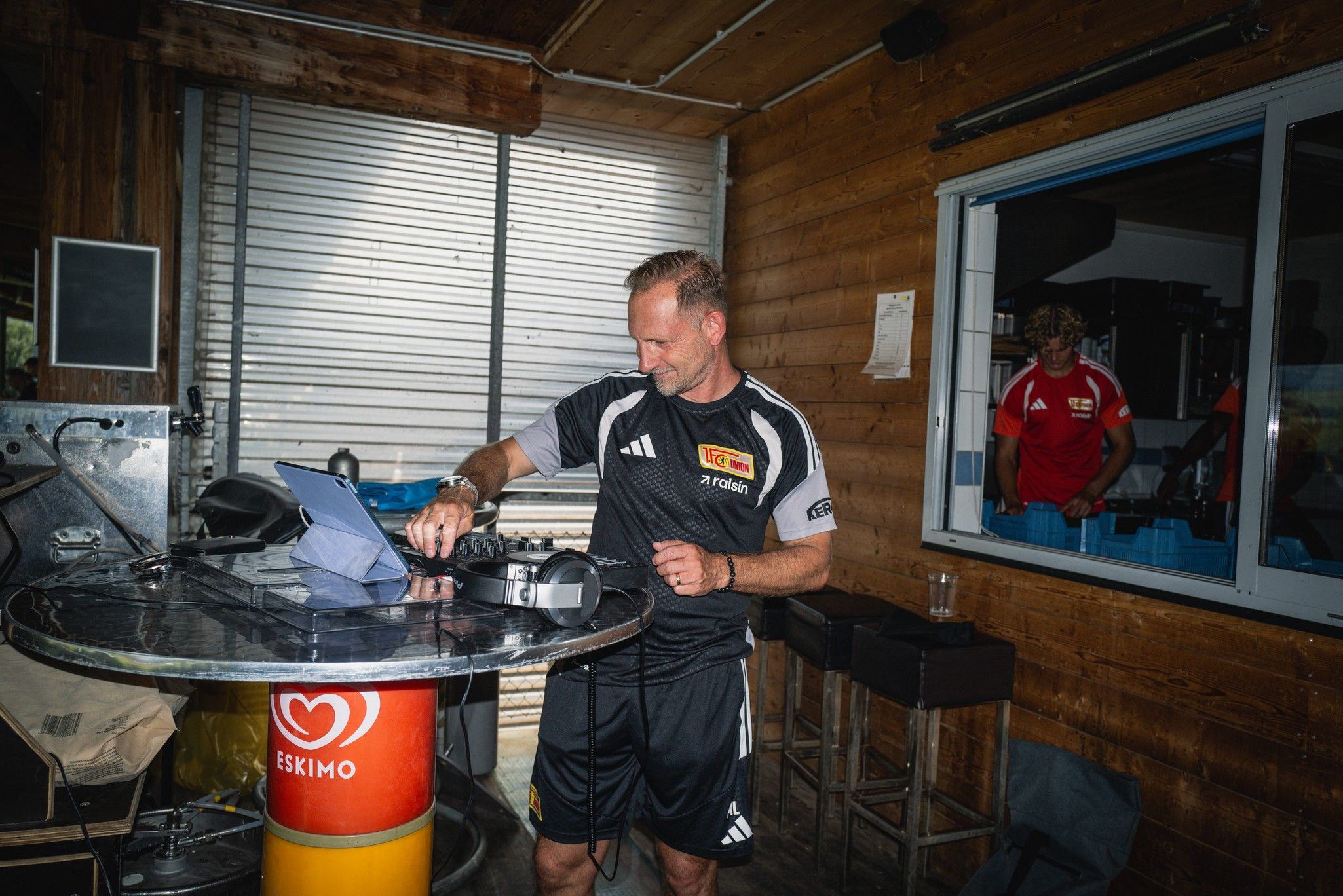 Ein Mann in Sportkleidung steht an einem Tisch mit einem Tablet und einer Kreditkartenmaschine in einem holzverkleideten Raum.