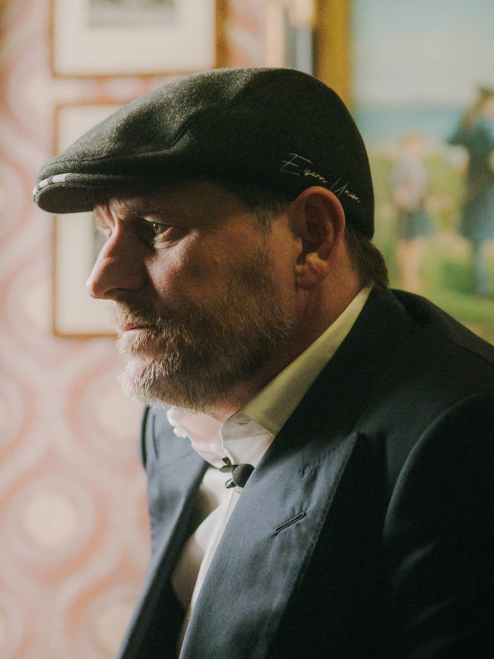 Ein Mann mit Bart und Mütze, der in einem eleganten Anzug gekleidet ist, schaut nachdenklich zur Seite vor einem dekorativen Hintergrund.