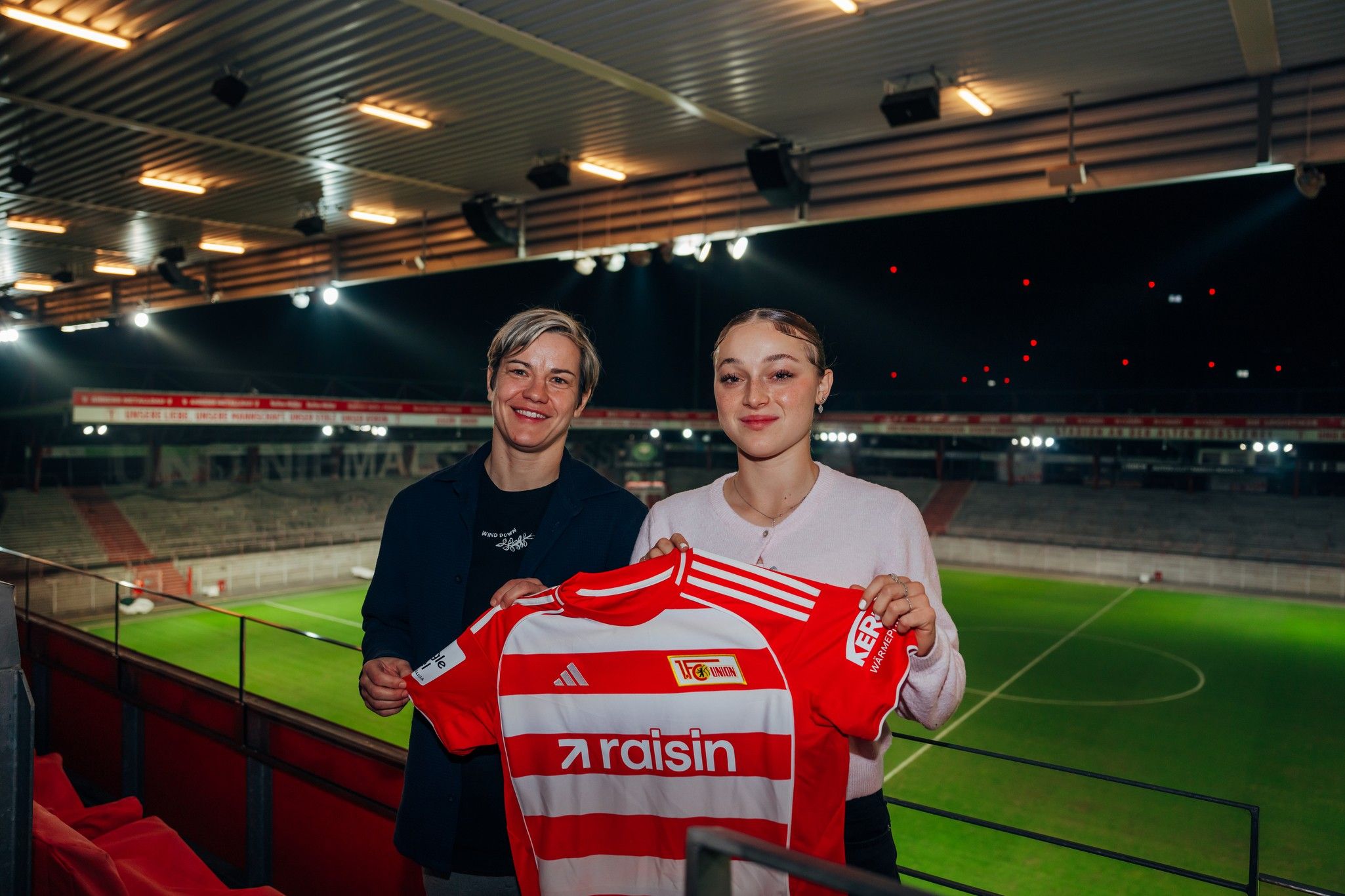 Zwei Personen halten ein Fußballtrikot mit dem Logo "raisin" im Stadion in der Hand. Im Hintergrund ist das Spielfeld zu sehen.