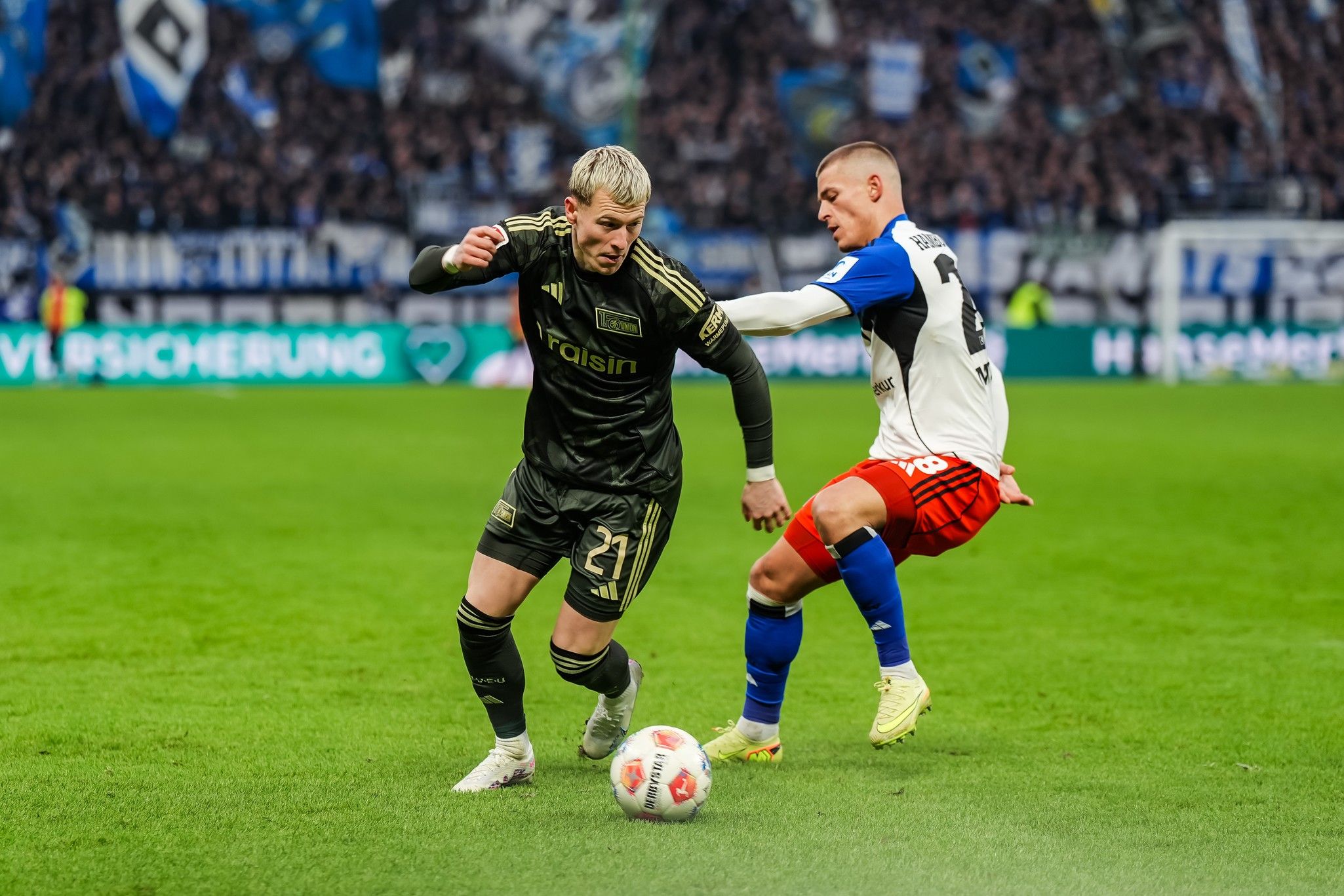 Ein Fußballspieler in dunkler Ausrüstung dribbelt den Ball, während ein gegnerischer Spieler in heller Ausrüstung ihn verfolgt.