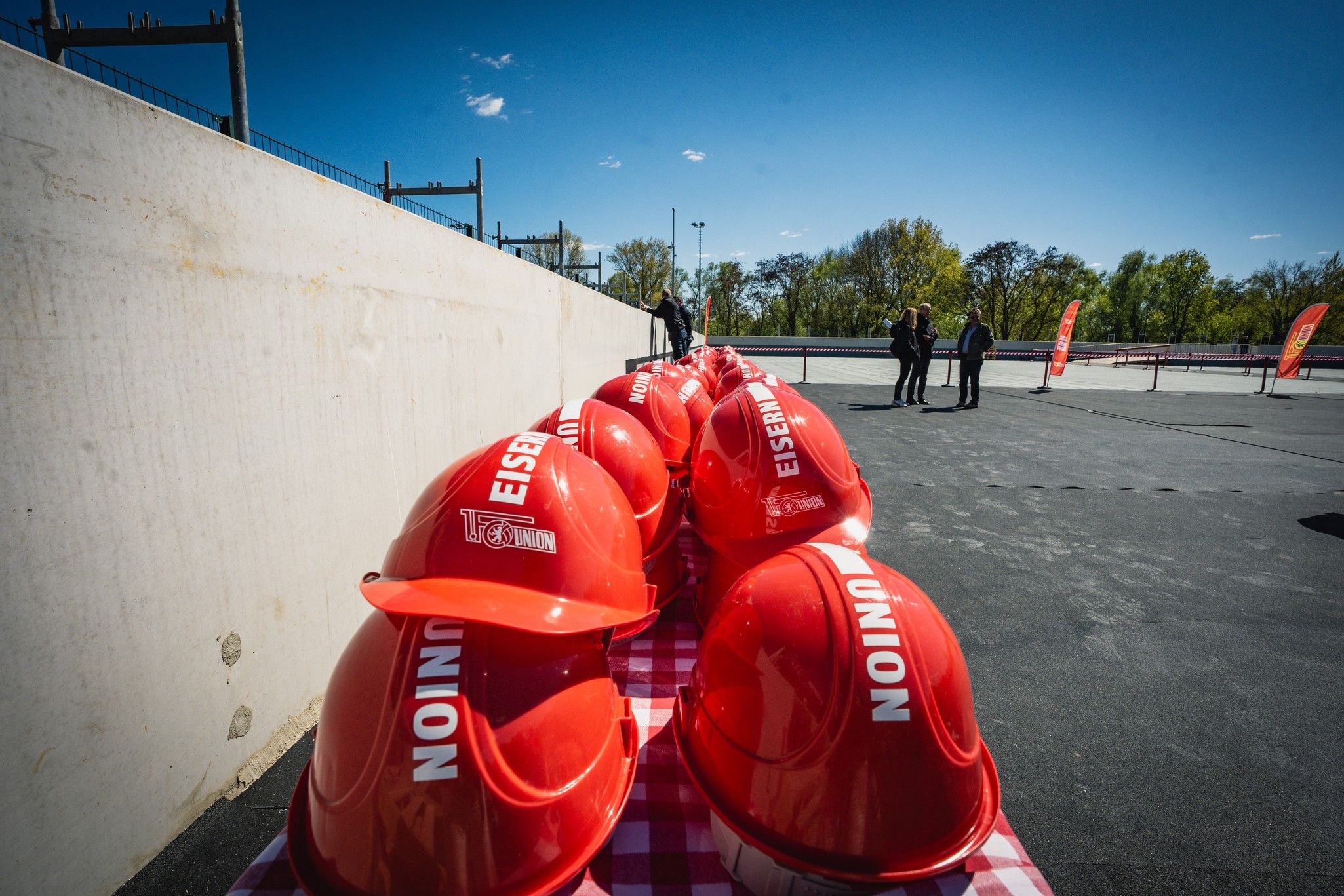 Roten Bauhelme stehen auf einem Tisch, im Hintergrund sind Menschen und Bäume sichtbar, bei klarem, blue Himmel.