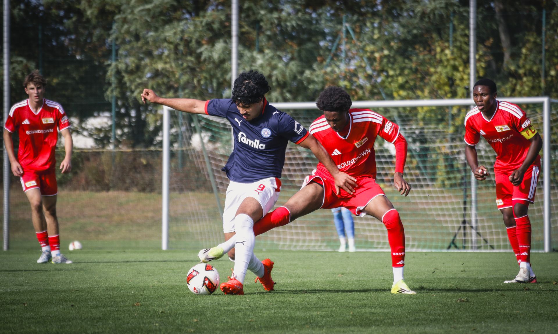 Ein Fußballspieler in blauer Uniform kämpft um den Ball gegen einen gegnerischen Spieler in roter Uniform auf einem grünen Rasenfeld.