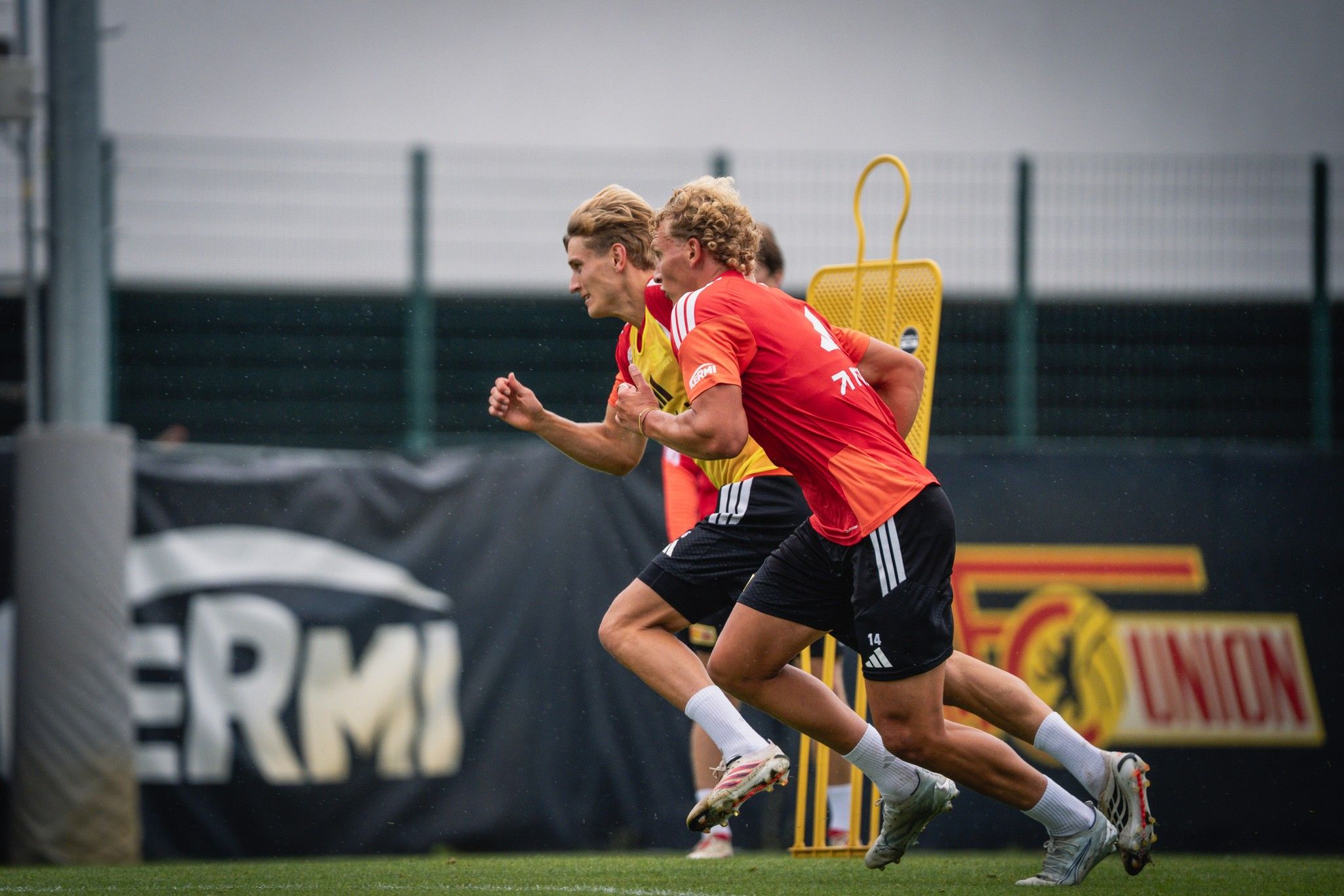 Zwei Fußballspieler in Trainingsanzügen sprinten auf einem Sportfeld, während ein Trainingsgerät im Hintergrund steht.