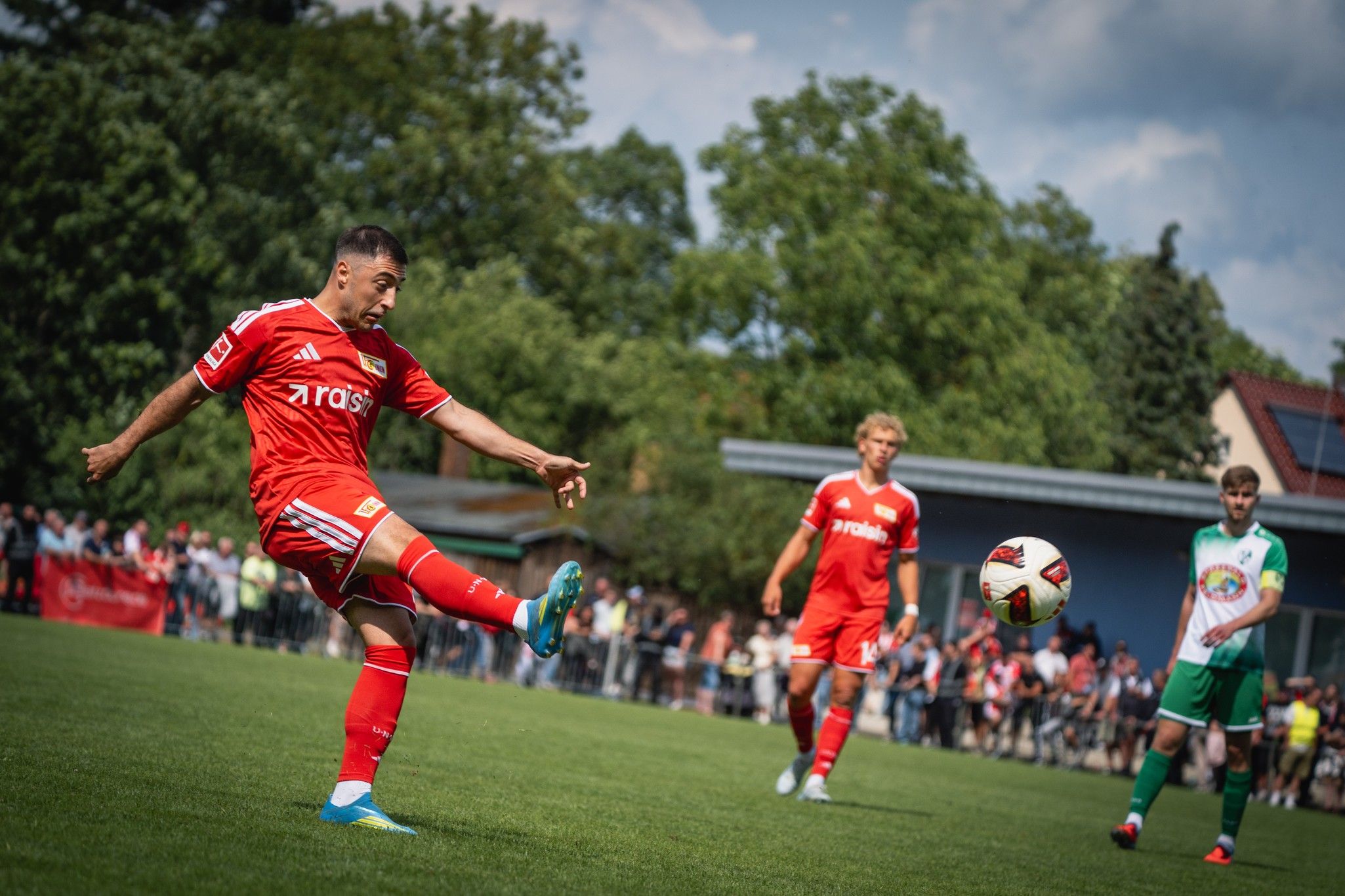 Ein Fußballspieler in roter Uniform tritt den Ball auf einem grünen Platz, während Zuschauer im Hintergrund stehen.