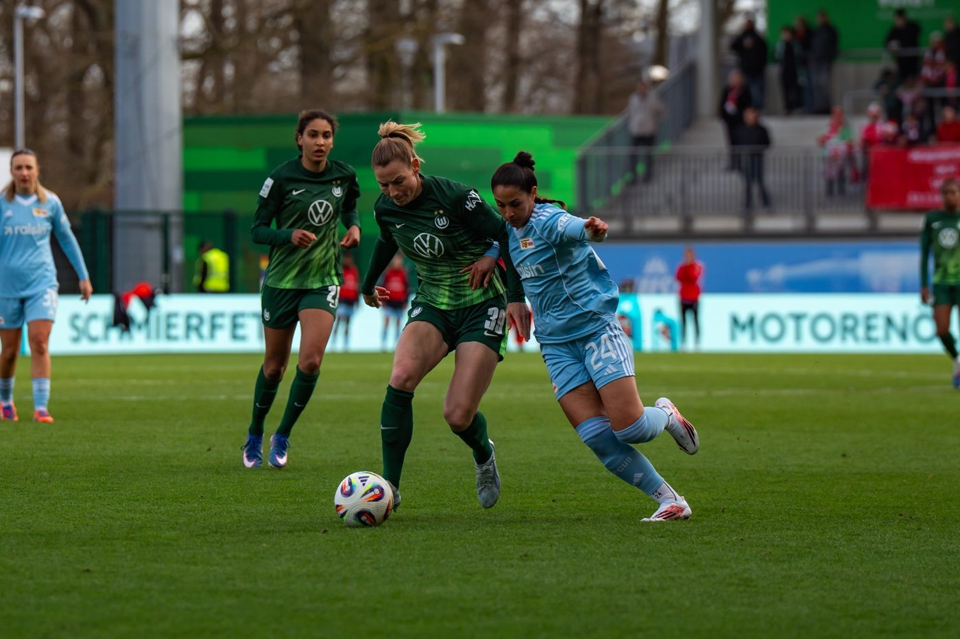 Eine Fußballspielszene mit zwei Spielerinnen im Wettkampf um den Ball, während die anderen Spielerinnen im Hintergrund stehen.