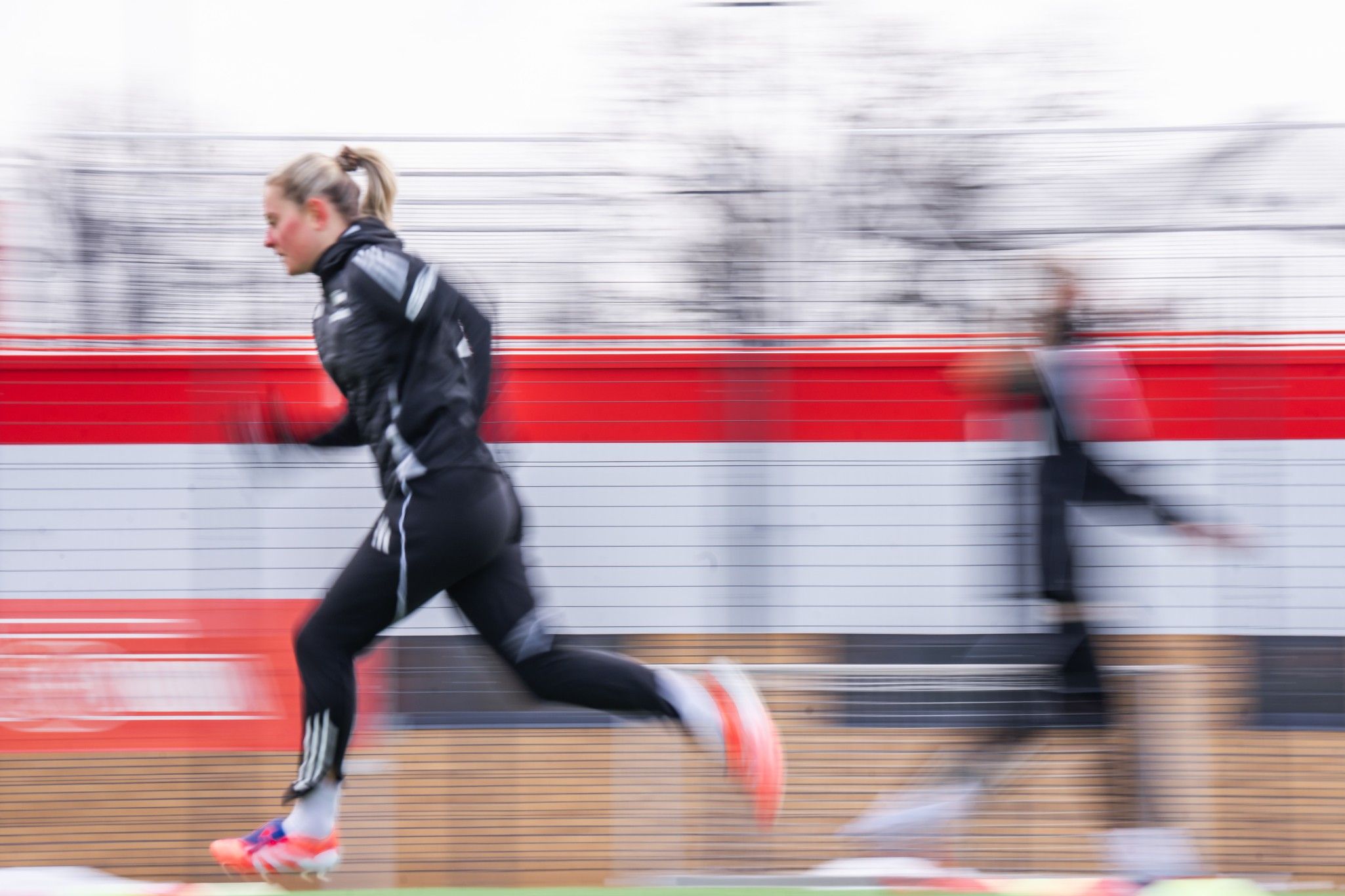 Läuferin in Sportkleidung sprintet schnell über den Platz, während eine weitere Person verschwommen im Hintergrund zu sehen ist.