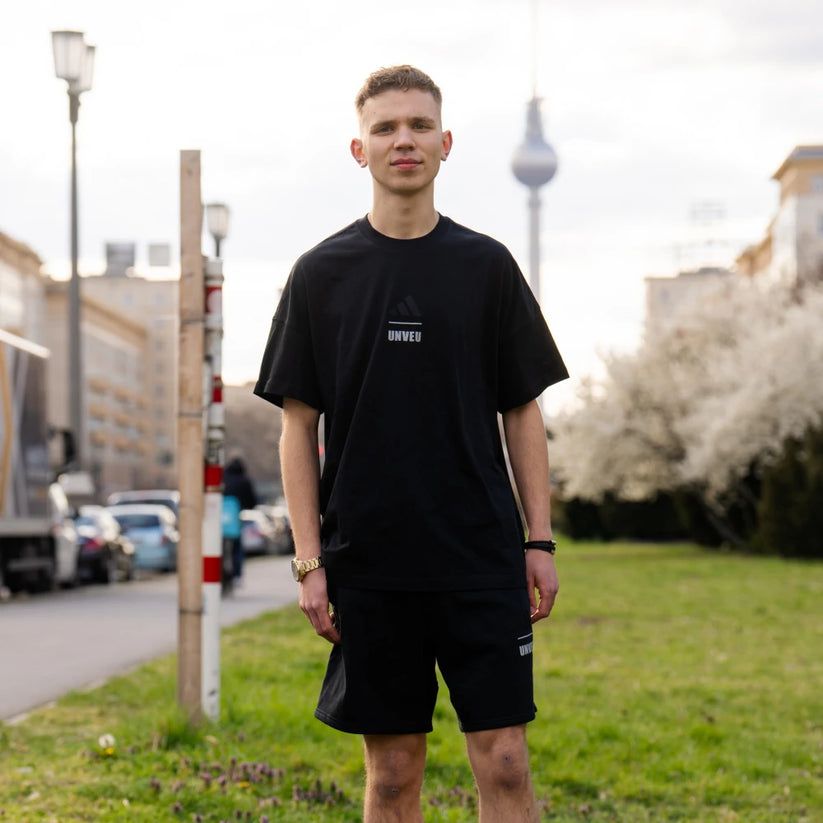 Mann in schwarzem T-Shirt und Shorts steht auf einer Wiese, im Hintergrund Gebäude und der Fernsehturm von Berlin.