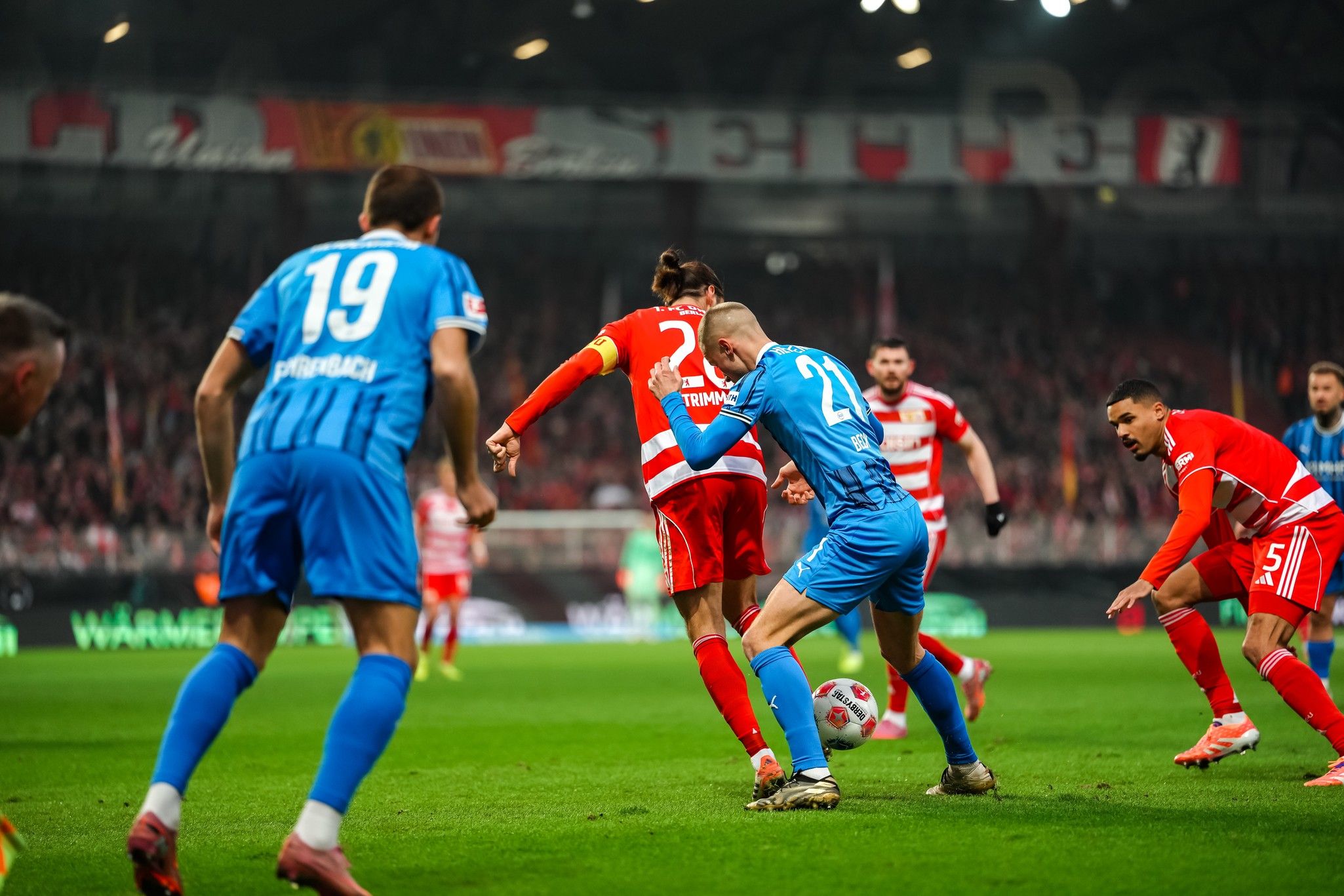 Fußballspiel zwischen zwei Mannschaften, Spieler in roten und blauen Trikots in Aktion auf dem Spielfeld mit Fans im Hintergrund.