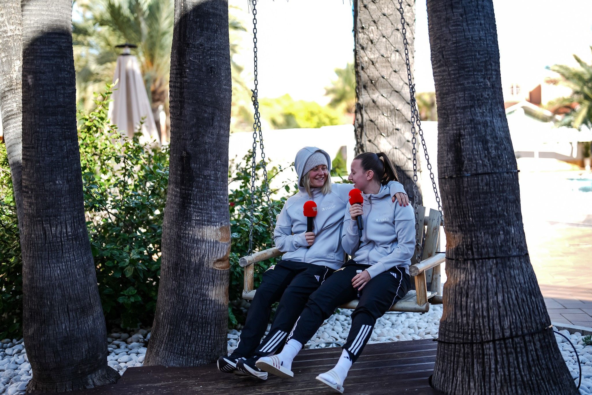 Zwei Personen sitzen auf einer Schaukel zwischen Palmen und halten Mikrofone. Beide tragen sportliche graue Jacken und schwarze Hosen.