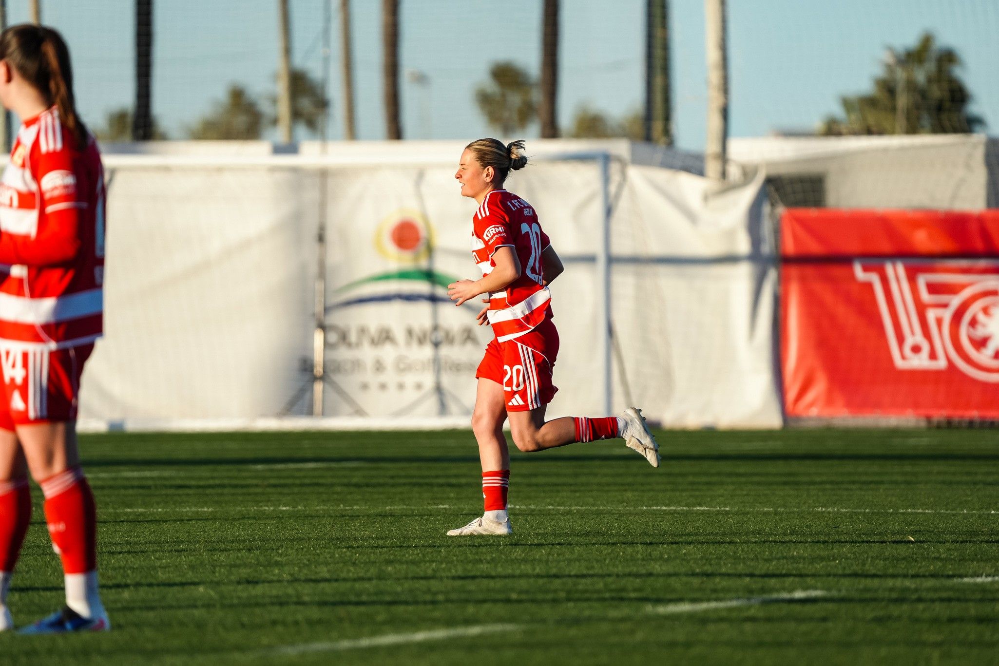 Eine weibliche Fußballspielerin in rot-weißer Uniform läuft über den Platz. Im Hintergrund sind Tore und eine Werbebande sichtbar.