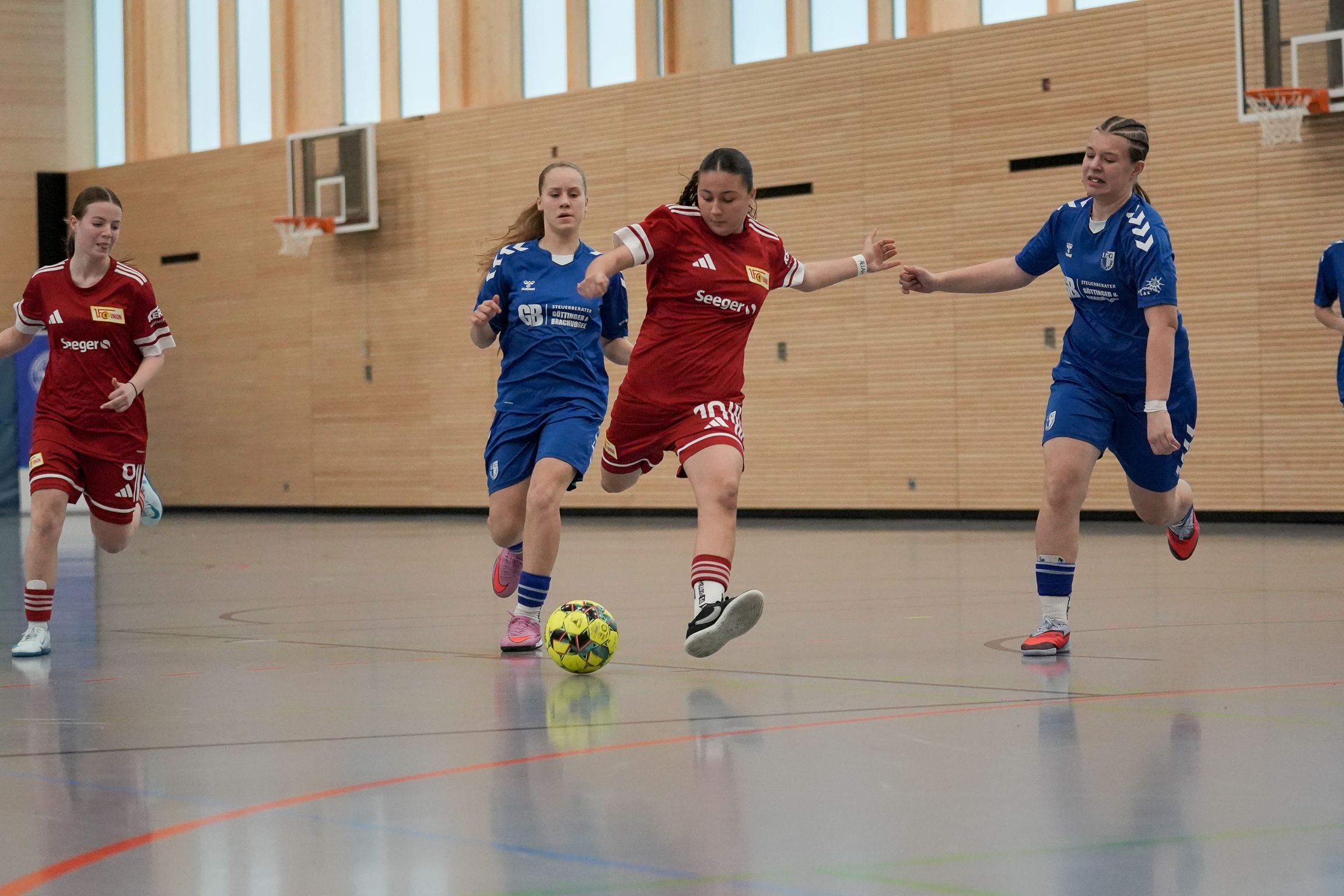 Eine Spielerin in rotem Trikot schießt einen Ball, während zwei Spielerinnen in blauen Trikots versuchen, ihn zu blockieren.