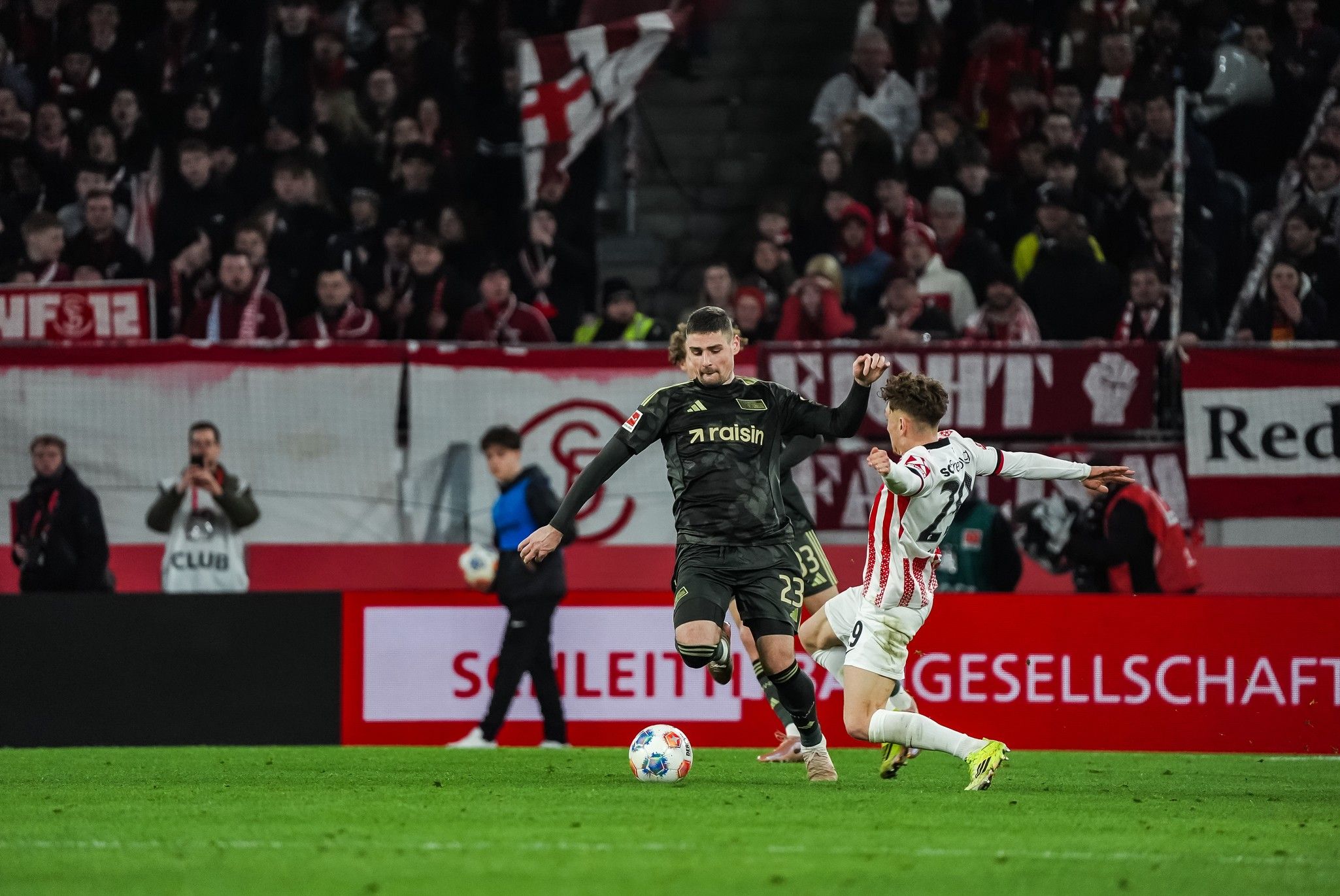 Ein Fußballspieler in schwarzer Kleidung dribbelt den Ball, während ein gegnerischer Spieler ihn von der Seite angreift. Zuschauer im Hintergrund.