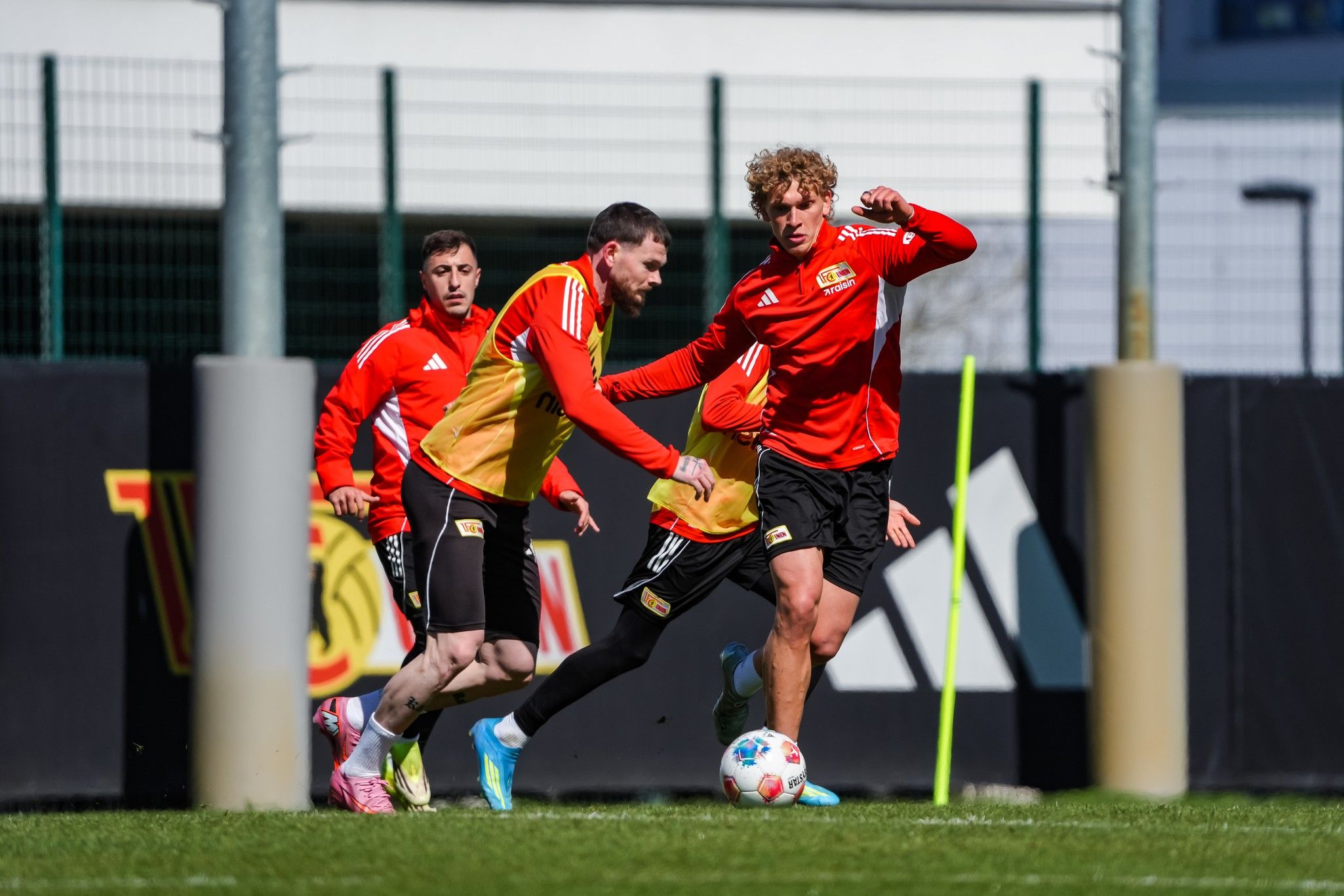 Drei Fußballspieler trainieren auf einem Rasenplatz, wobei zwei Spieler im intensiven Dribbling zu sehen sind.
