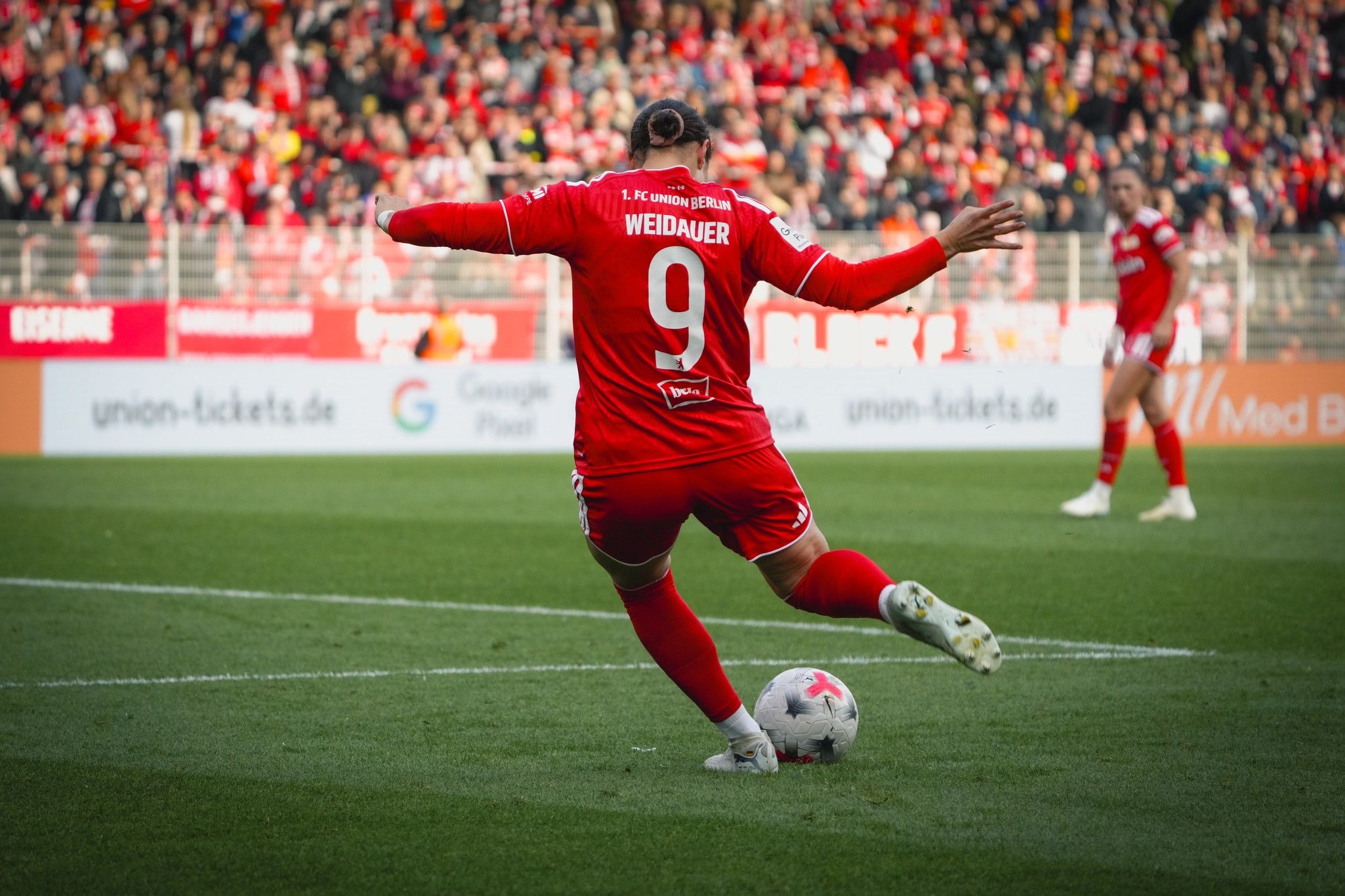 Ein Fußballspieler in roter Uniform tritt den Ball auf einem Stadionfeld mit jubelndem Publikum im Hintergrund.