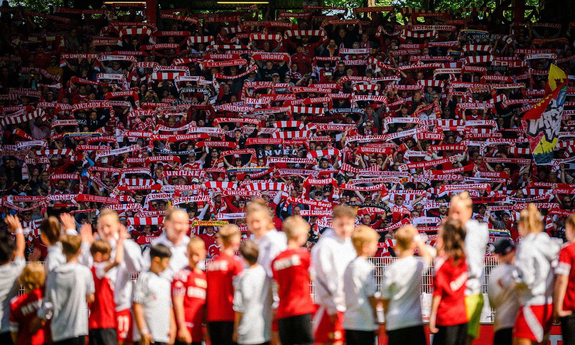 Eine große Menschenmenge im Stadion hält rote und weiße Schals hoch, während Mannschaften auf dem Spielfeld stehen.
