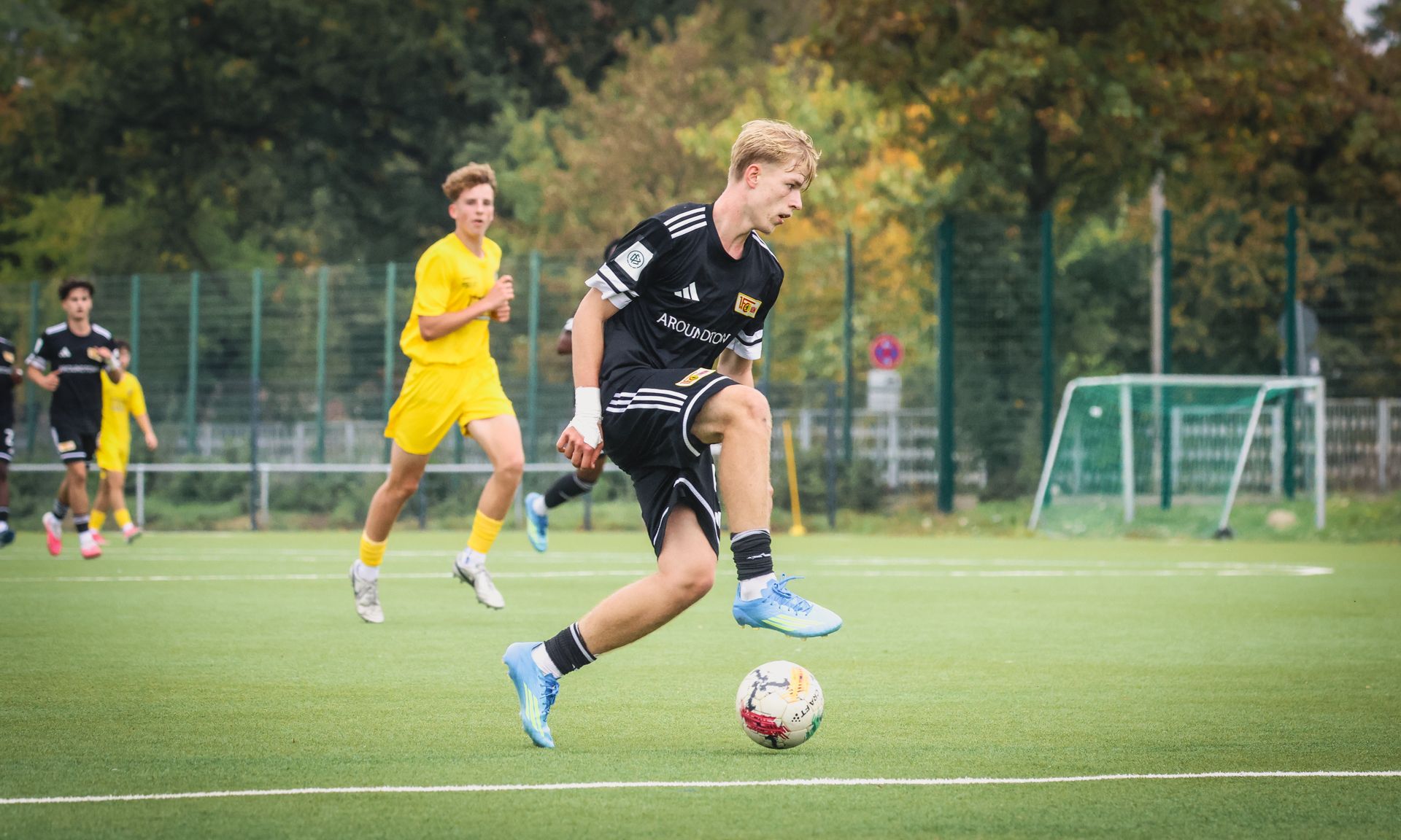 Ein junger Fußballspieler in schwarzer Kleidung dribbelt den Ball auf einem Kunstrasenfeld, während ein Gegner in Gelb im Hintergrund läuft.