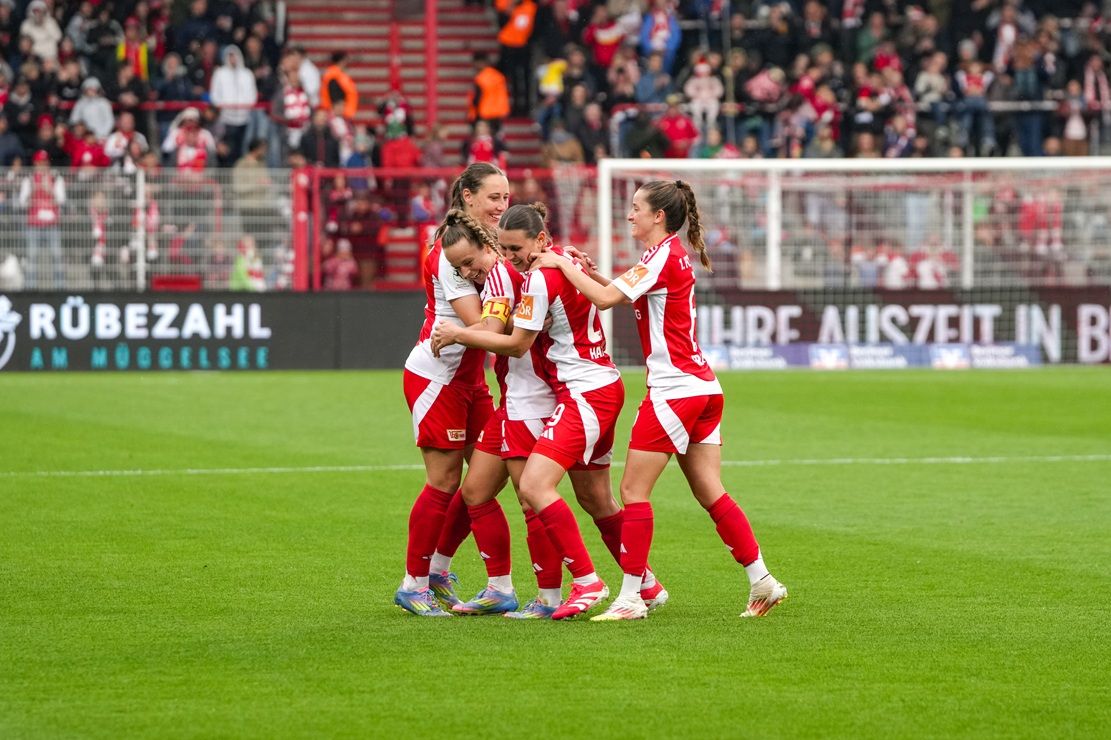 Frauenfußballmannschaft feiert ein Tor miteinander auf dem Spielfeld, mit jubelnden Fans im Hintergrund.