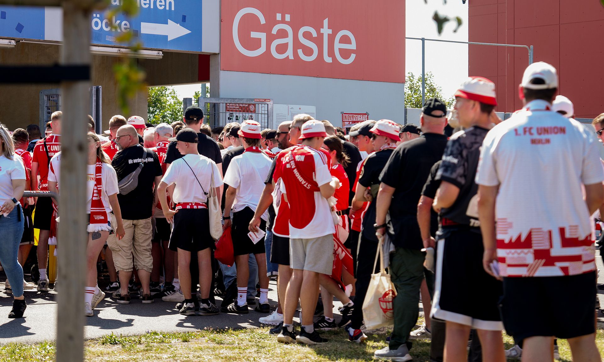 Menschenmenge in roten und weißen Trikots wartet an einem Eingang mit dem Schild "Gäste".