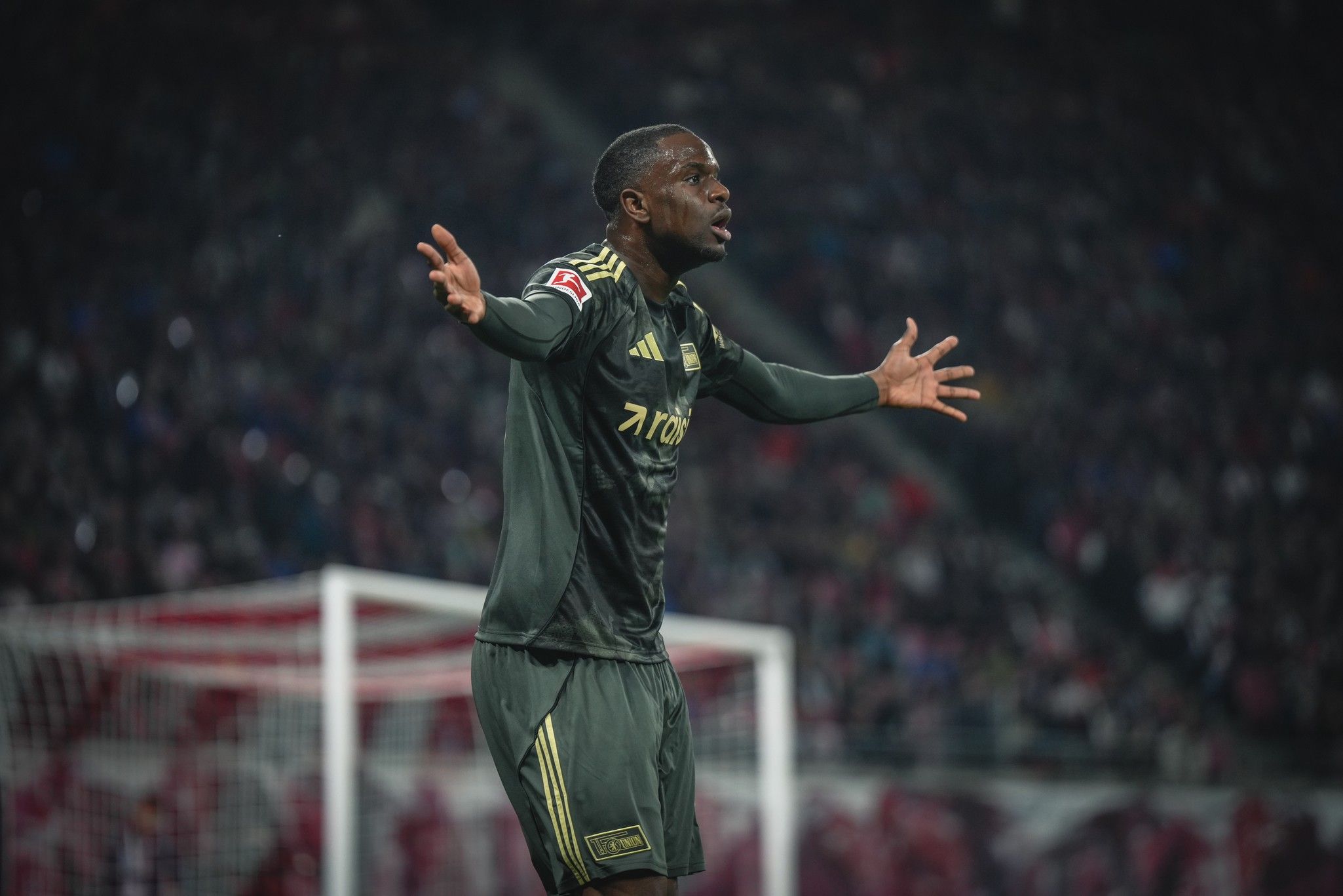 Ein Torwart in grauer Sportausrüstung steht auf dem Spielfeld und gestikuliert während eines Fußballspiels.