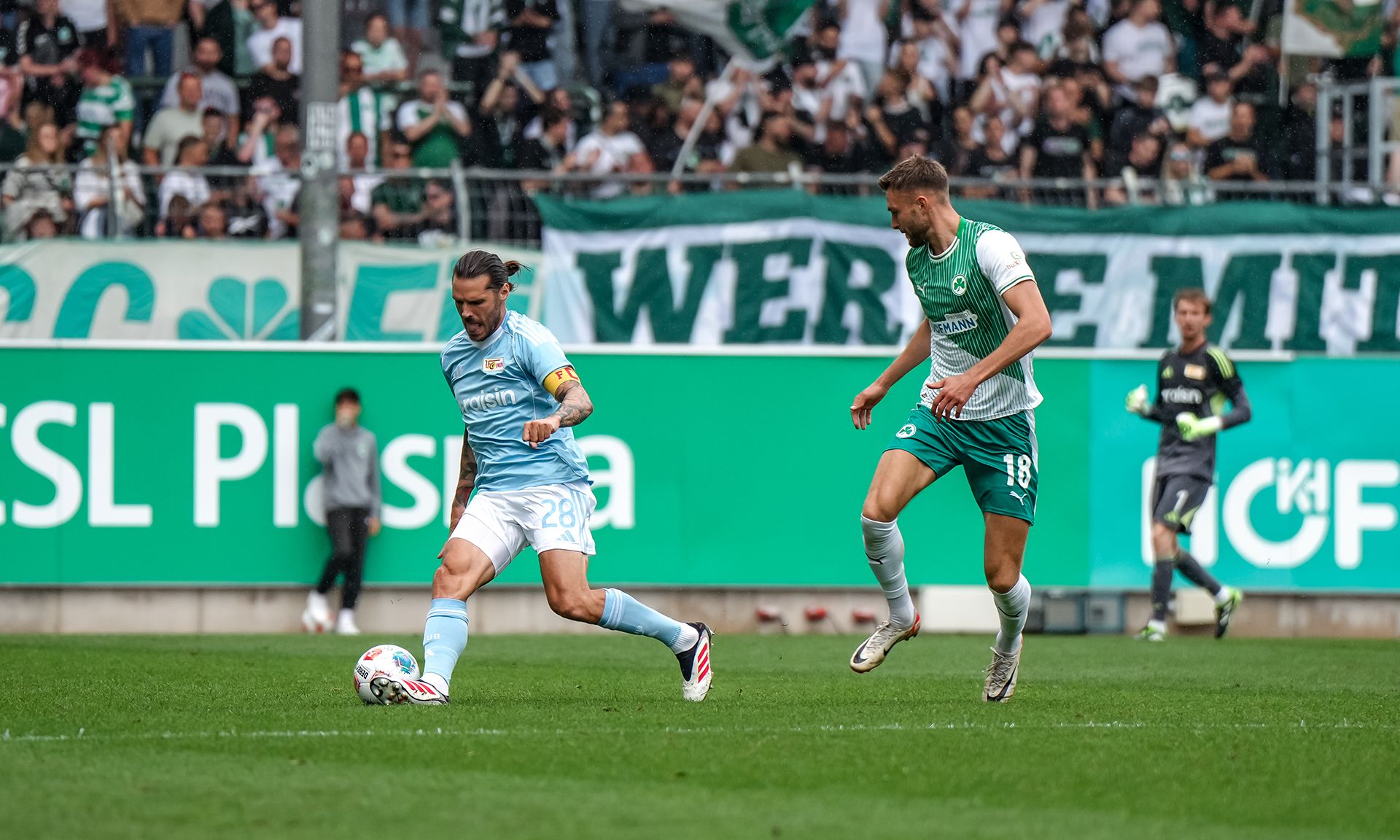 Ein Spieler in hellblauem Trikot passt den Ball, während ein gegnerischer Spieler in einem weißen Trikot ihn verfolgt.